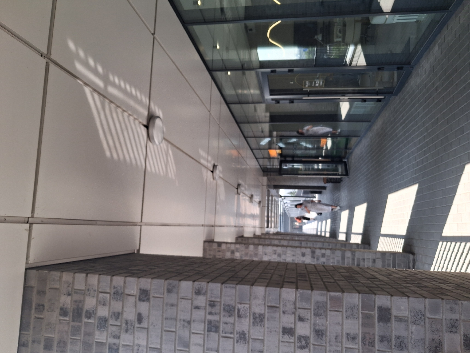 This image depicts a modern, covered walkway in Novosibirsk, Russia, likely adjacent to a commercial building. The walkway is paved with grey rectangular bricks, and the ceiling is constructed from white modular panels, with circular light fixtures attached. The left side of the walkway is bordered by a robust, grey brick wall. The right side features a continuous glass facade of a building, reflecting the surrounding environment and the interior of the establishments. Several individuals are visible in the distance, walking along the walkway. One woman in a light-colored dress is prominent, walking towards the viewer. Another person in an orange shirt is also visible further down the path. The sun casts distinct striped shadows across the walkway from an unseen overhead structure, suggesting it is a bright, sunny day. The overall impression is one of a well-maintained urban passage, blending architectural elements with natural light.