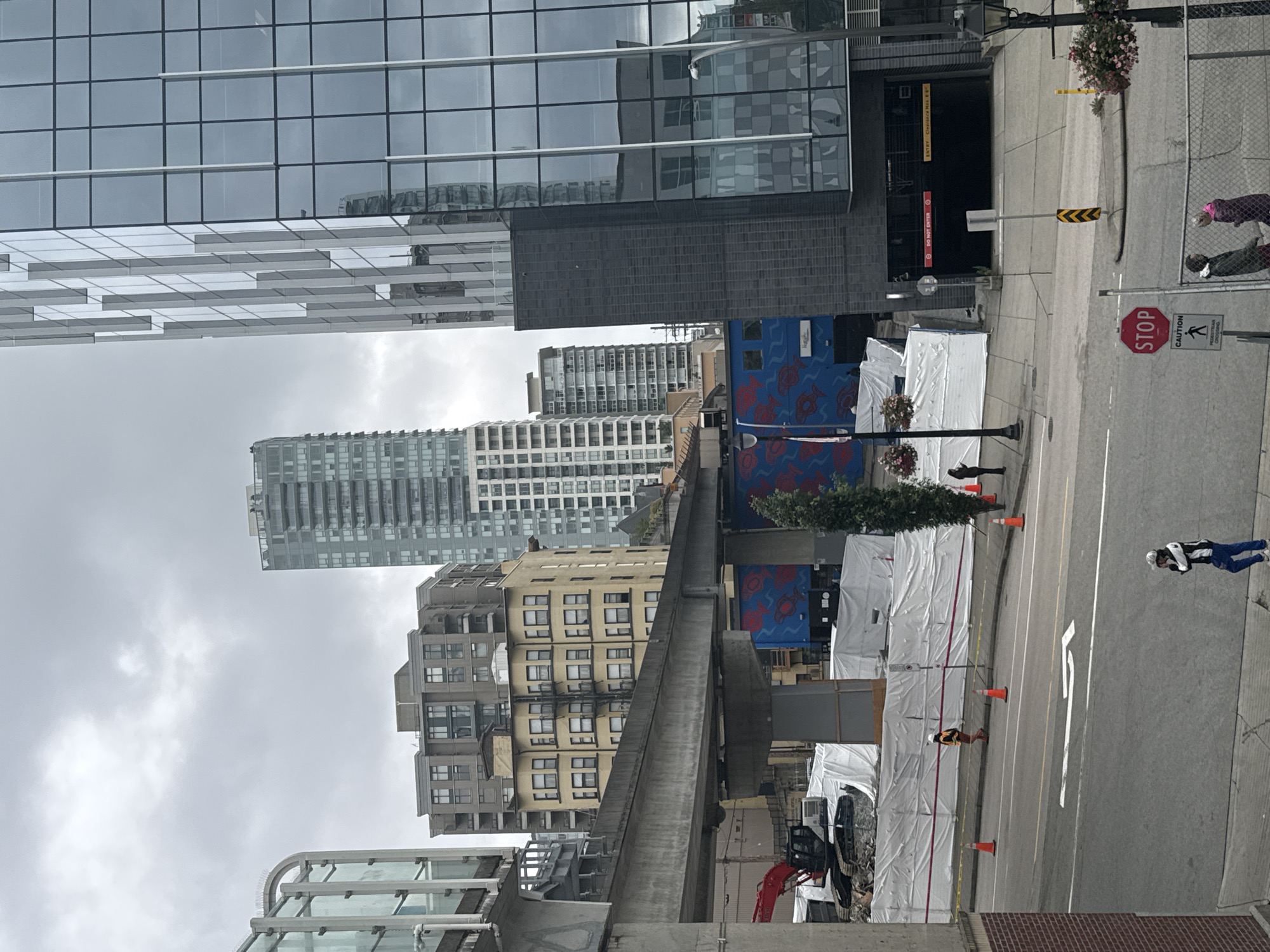 The image depicts an overcast day in a city, likely New Westminster, Canada, with a focus on urban infrastructure and ongoing construction. In the foreground, a street runs horizontally, with a pedestrian walking on the sidewalk to the right, wearing a white hard hat, dark jacket, and blue pants, appearing to be on their phone. To the left, a brick building borders the sidewalk. Across the street, a construction site is visible, partially covered with white tarps and surrounded by orange traffic cones. An excavator is present at the site. Elevated above the construction is a concrete overpass, part of a transit system.
Beyond the construction, several buildings rise into the sky. To the left of the overpass is a multi-story building with a glass facade. Further back, a lighter-colored, older apartment building with many windows stands out. In the center, a tall, slender skyscraper with a patterned glass exterior dominates the skyline, reflecting the cloudy sky. To the right of this skyscraper, another tall building with a dark facade and grid-like windows is visible, partially obscuring a dark entrance to an underground parking garage. A red sign reading "DO NOT ENTER" and a yellow sign indicating "ENTRY CLEARANCE MAX. 6' 6"" are affixed above the garage entrance.
A large blue mural with red fish-like imagery is visible on the side of a building behind the construction site. A lone tree stands near the mural. In the background, the sky is heavy with clouds. Two individuals are standing near a chain-link fence to the right of the stop sign in the foreground, appearing to be in conversation.