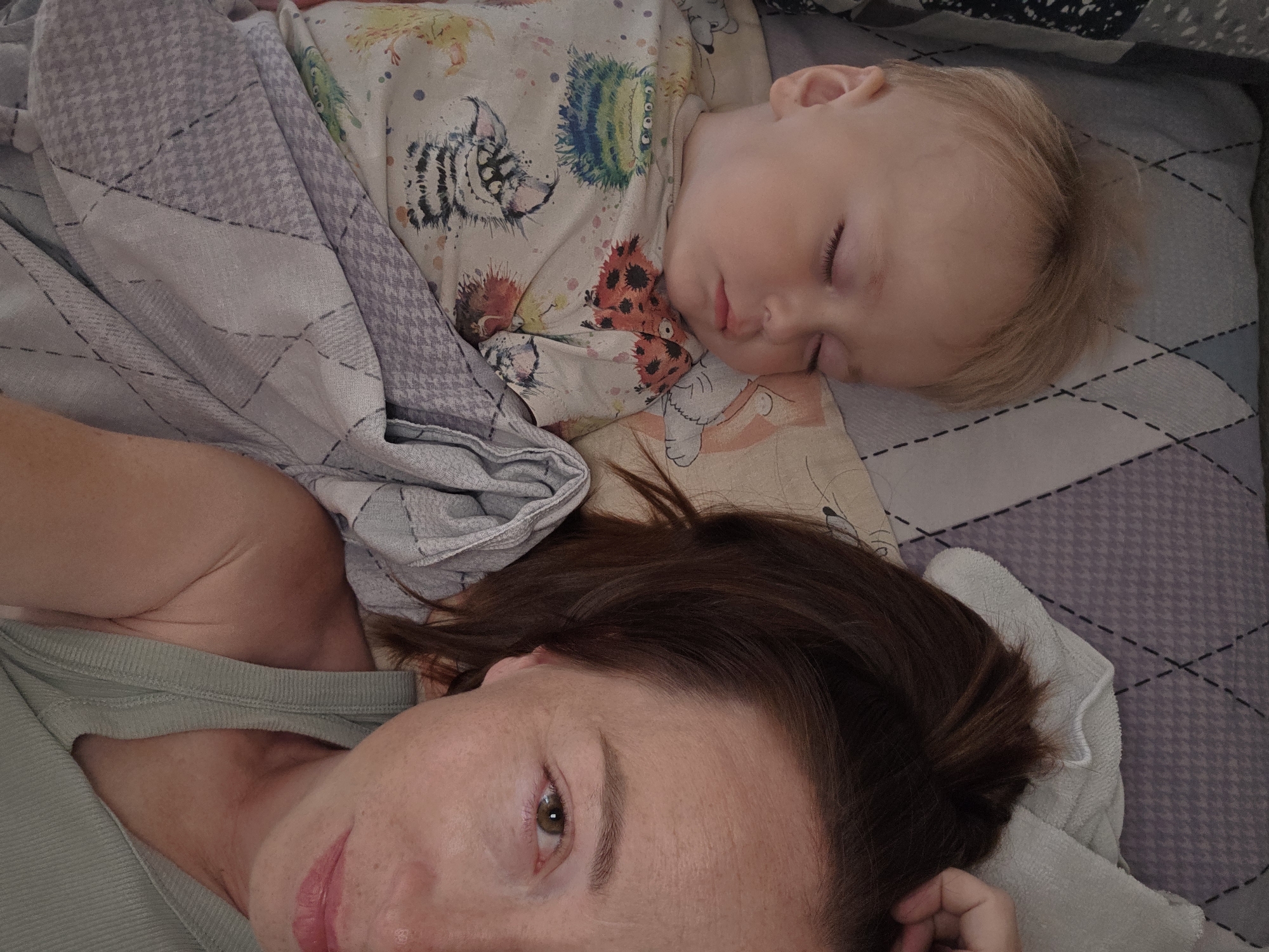 A young child with blonde hair is sleeping on their back, covered by a grey and white patterned blanket. The child is wearing a light-colored onesie with a colorful monster print. Next to the child, a woman is lying down, her head resting on a pillow. Her eyes are open and she is looking towards the camera. Her dark brown hair is pulled back into a ponytail. The background shows a bed with patterned sheets. The scene is indoors. The image was taken in Chaikovsky, Russia.