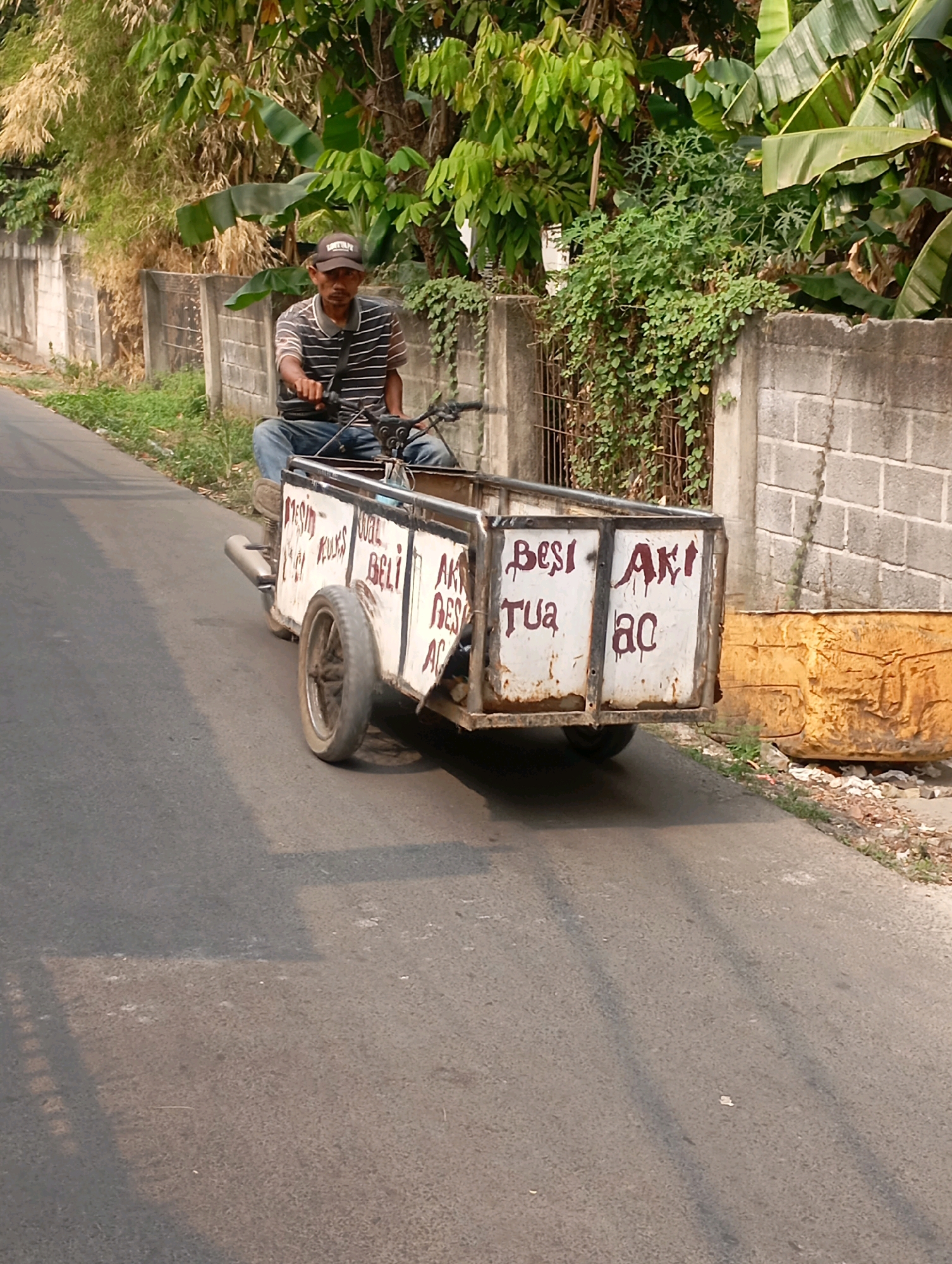 A man wearing a striped shirt and jeans rides a three-wheeled vehicle with a trailer on a paved road in Babelan, Indonesia. The trailer is white with red lettering that appears to say "MESIN KULK BELI AKI TUA AC". The man is wearing a dark baseball cap. Behind him, a concrete wall runs along the side of the road, with lush green foliage and banana trees growing behind it. To the right of the road, a large, weathered yellow tank sits next to a pile of debris. The scene suggests a commercial or recycling operation.