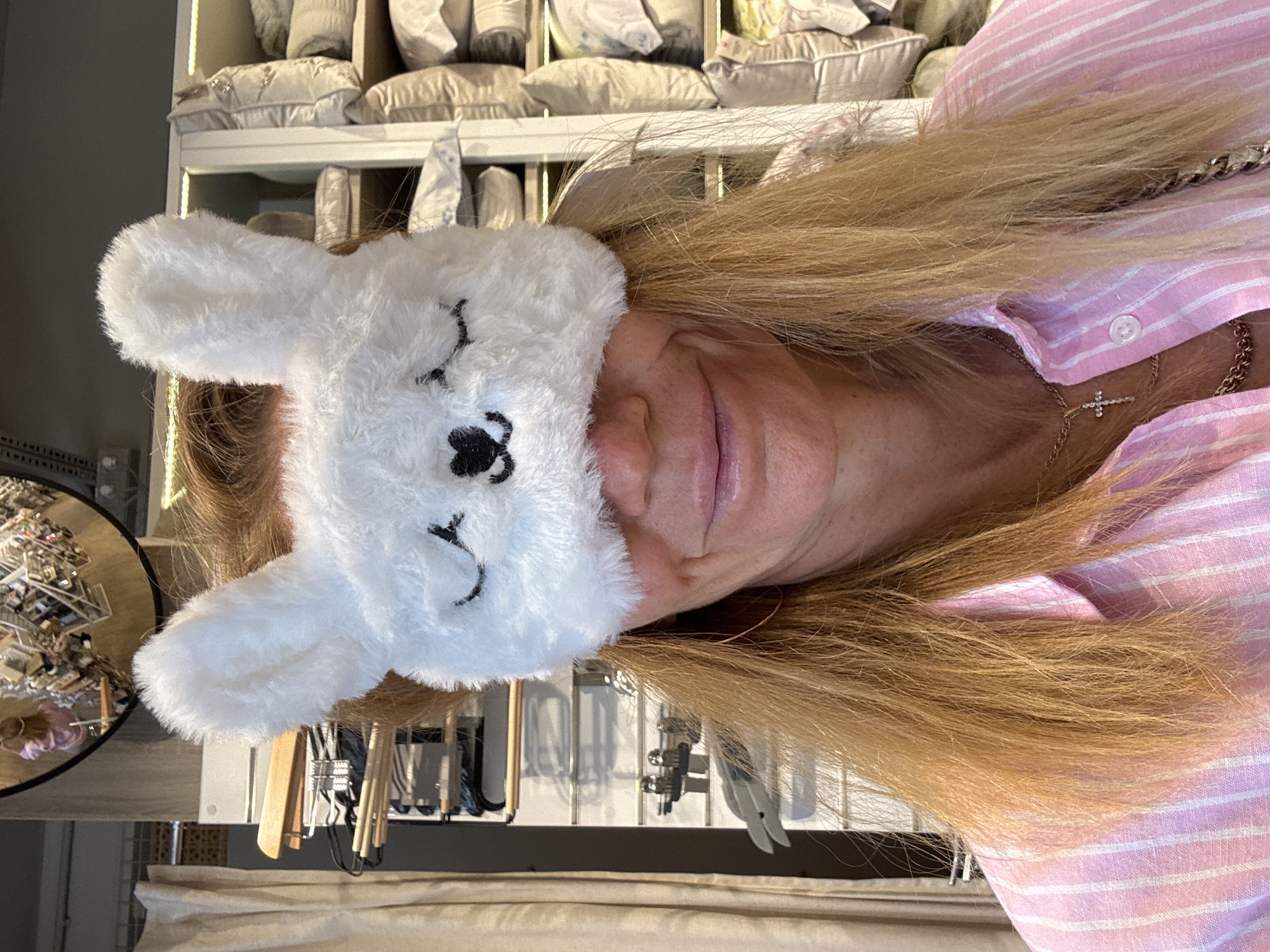 A fair-skinned person with blonde hair wears a white, plush rabbit eye mask. The person is wearing a pink and white striped shirt and a gold necklace with a cross pendant. The background shows a shelf with pillows, a mirror with items displayed on it, clothing racks with hangers, and neutral-colored fabric. The location is not discernible from the image.