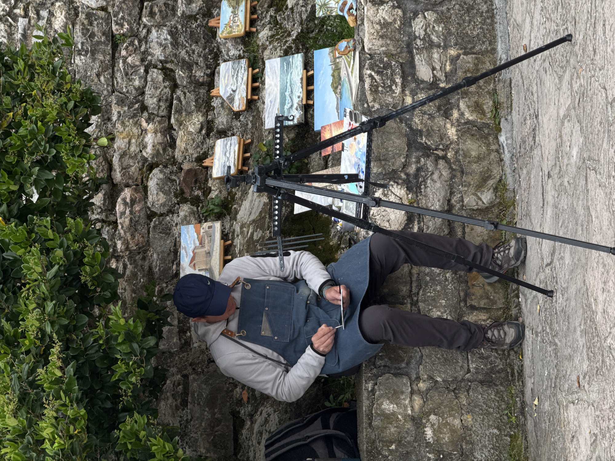 An artist sits on a stone ledge, seemingly in the city of Budva, Montenegro, painting with a brush. He wears a blue baseball cap, gray hoodie, dark-colored pants, and an apron. Paintings are displayed on the stone wall behind him, held by small easels. A black tripod stands next to the artist. Green foliage is present on the left side of the image. The ground and the wall are made of rough, light-colored stone.