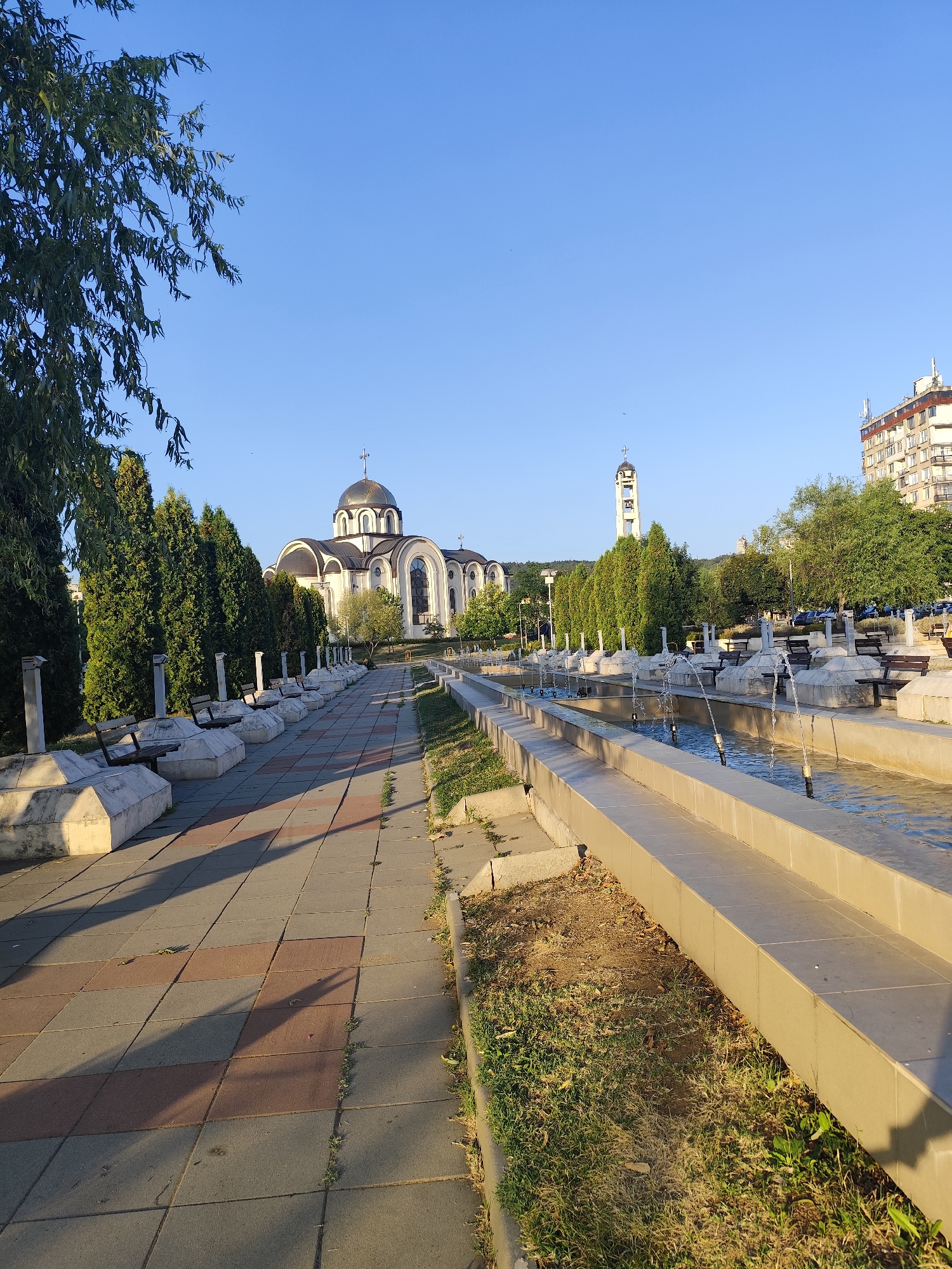 The image captures a view of the central pedestrian zone in Montana, Bulgaria, characterized by a prominent Orthodox church with a golden dome and a tall bell tower. A linear fountain with multiple water jets runs parallel to the walkway, which is paved with square tiles of red and gray. Lined along the walkway are benches with stone bases, interspersed with decorative concrete structures and cypress trees. The sky is clear and blue, indicating a sunny day. A modern apartment building is visible in the background, to the right of the church. No people are present in the scene. The lighting suggests it is either early morning or late afternoon, given the long shadows cast by the trees and benches.