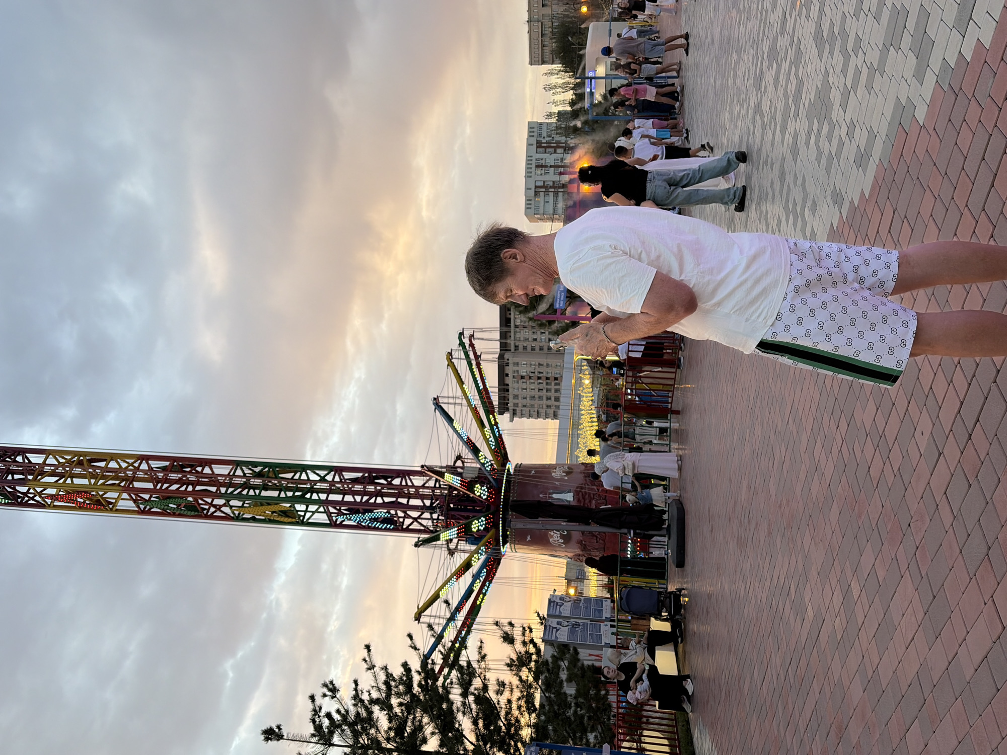 A man in a white t-shirt and patterned shorts stands in an amusement park, looking at his phone. He is in the foreground, facing away from the camera and towards the right. Behind him, a tall, colorful amusement ride with lights is visible, along with other park-goers walking around. The sky is cloudy with hints of sunset colors. Buildings are visible in the background. The ground is paved with red and grey bricks.