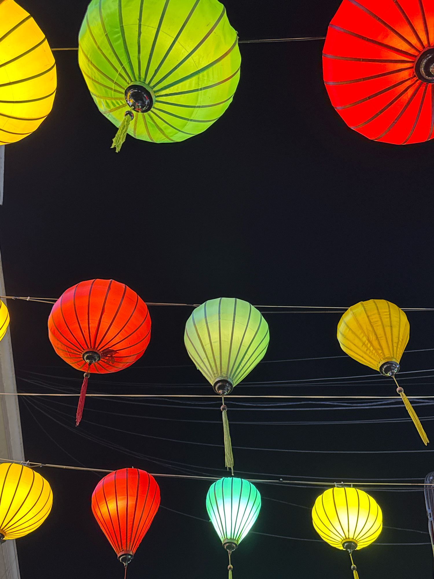 The image displays a collection of vibrantly colored lanterns suspended against a dark night sky. The lanterns are of various sizes and colors, including yellow, green, and red, and feature a ribbed, spherical design. Some lanterns have decorative tassels hanging from them. They are strung across multiple wires, suggesting an outdoor setting, possibly a street or market. The lighting from the lanterns creates a warm and festive atmosphere. The specific location in Nha Trang, Vietnam, is not discernible from the image itself, as the scene focuses solely on the lanterns and the dark sky. There are no people or any other discernible objects present in the scene.