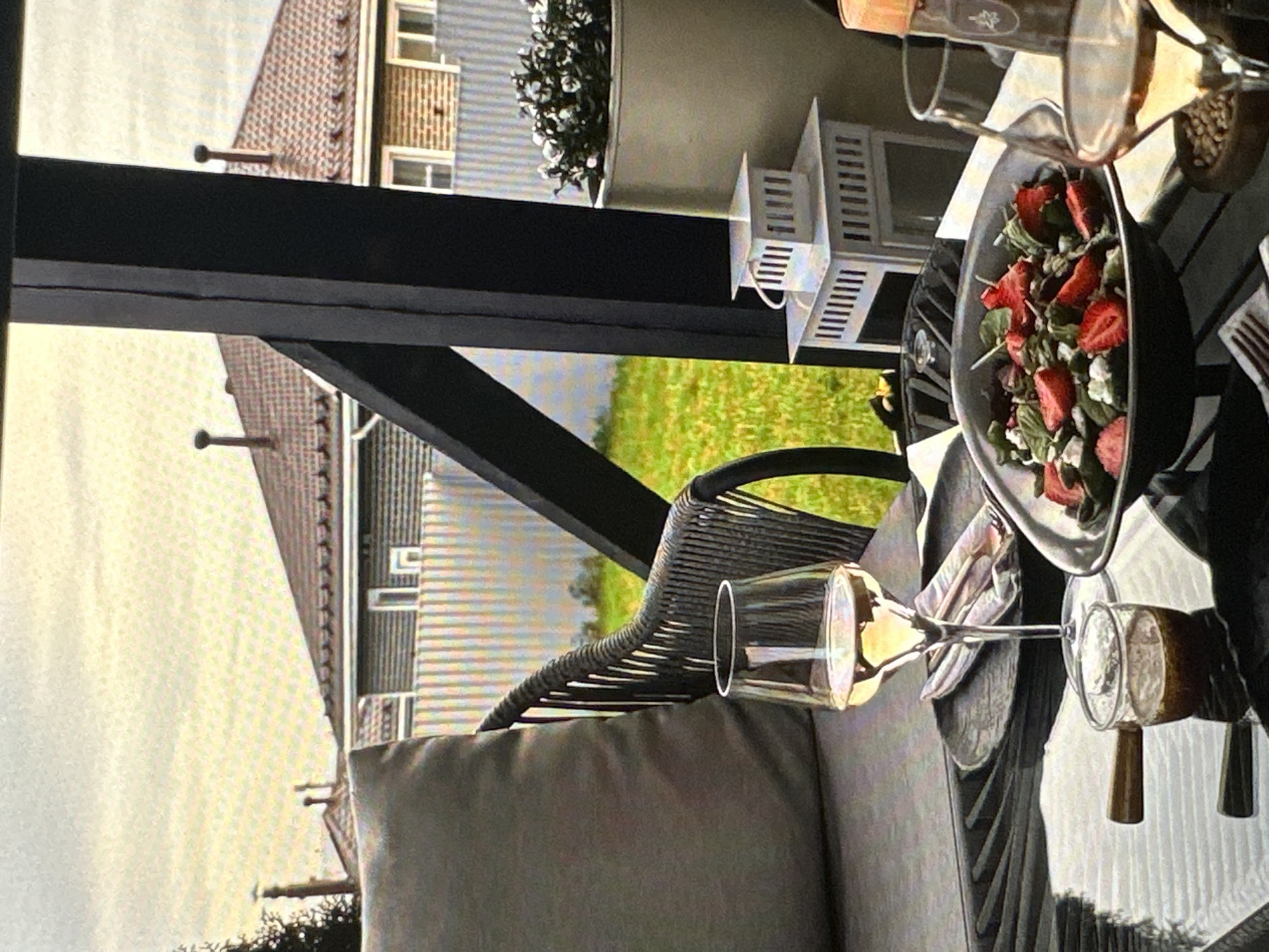 A partially rotated, medium-shot image depicts a dining area, presumably on a patio or balcony. A black, angular structure, possibly part of a pergola or architectural feature, frames the view towards a residential area and greenery. Visible buildings feature light-colored facades and pitched roofs. In the foreground, a cushioned patio set provides seating. A table is set with a salad of mixed greens and sliced strawberries served in a dark bowl, along with wine glasses. An ornamental lantern and potted plants are placed on the table. The composition suggests an outdoor dining setting in a residential area. The location is unknown but is not Yerevan, Armenia.