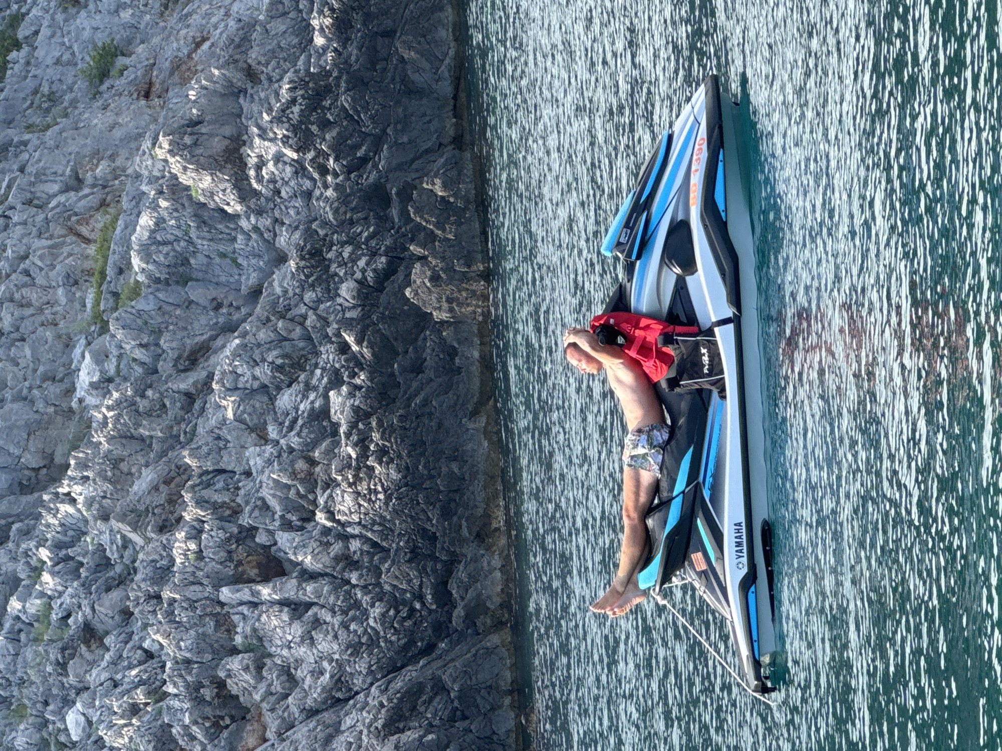 A man relaxes on a jet ski docked next to a rocky cliff face. The man is lying on his back with his head resting on a red life vest. He is wearing patterned swim trunks. The jet ski is grey and blue with "YAMAHA" visible on the side. The water is a deep blue-green with ripples reflecting sunlight. The rocky cliff is grey and textured, with small patches of greenery. The scene suggests a sunny day in a coastal location. The time of day appears to be daytime due to the bright sunlight and clear visibility. The location is identified as Dubraljevina, Montenegro.