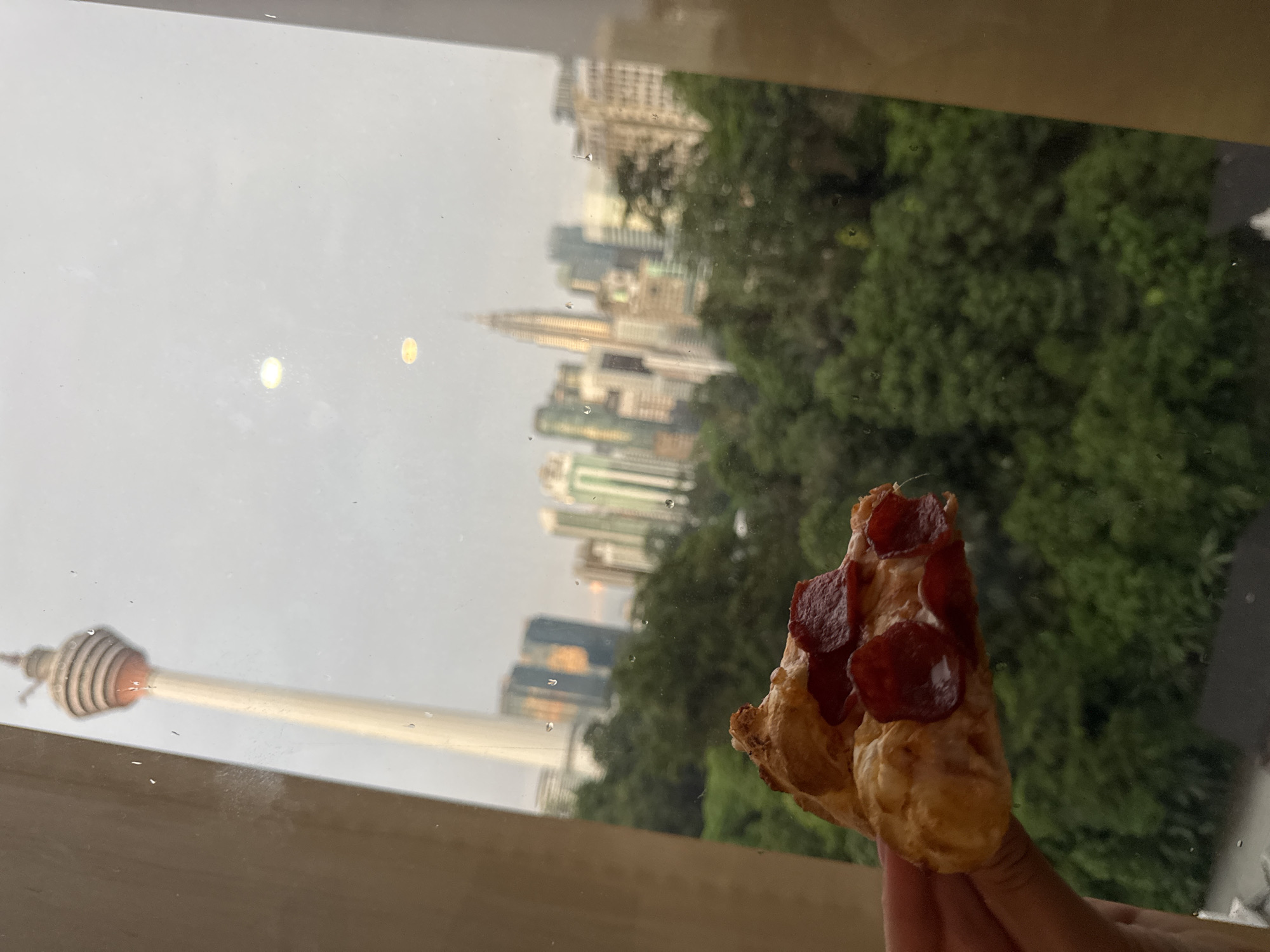 The image presents a vertical outdoor view of Kuala Lumpur, Malaysia, seen through a rain-splattered window. In the foreground, a hand holds a piece of food resembling a mini pepperoni pizza or a savory pastry topped with several slices of pepperoni. The windowpane is covered with numerous water droplets, indicating it is raining or has recently rained.
Beyond the window, the city skyline is visible but out of sharp focus. The prominent Menara Kuala Lumpur (KL Tower) stands distinctively on the left side of the frame, a tall structure with a unique observation deck design. In the mid-ground and further distance, numerous skyscrapers contribute to the urban landscape, with the iconic Petronas Twin Towers faintly discernible among them. A significant amount of lush green foliage and trees is visible, situated between the foreground and the distant buildings, suggesting a park or green space within the city.
The sky is bright but uniformly grey and overcast, indicating a daytime setting without direct sunlight. The scene conveys a quiet, contemplative moment of enjoying a snack while observing the bustling city from a sheltered vantage point. There is no visible text in the image.