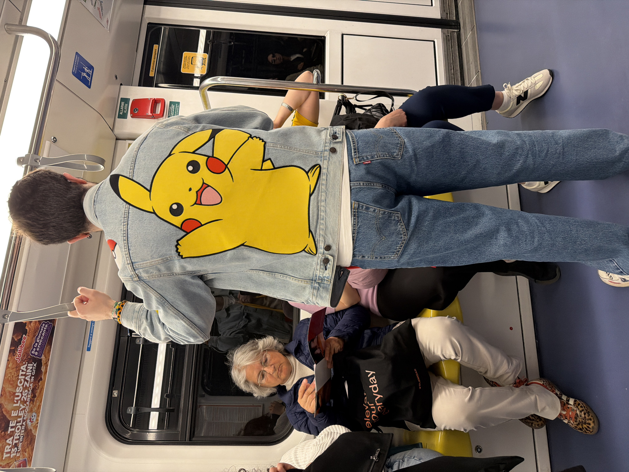 The image shows the interior of a public transport vehicle, likely a metro or train in Milan, Italy, filled with passengers. The perspective is from behind a young person standing in the foreground, whose back is to the camera. This individual wears a light-washed denim jacket featuring a large, vibrant yellow Pikachu character on the back, paired with blue denim jeans. They have short dark hair with an earring and are holding onto a grey grab strap.
Further into the carriage, several other passengers are visible. One person is seated, wearing dark pants and white New Balance sneakers with a distinctive 'N' logo. An older woman with grey hair and glasses is seated in the mid-ground, engrossed in reading a book or magazine. She is dressed in a dark blue padded jacket over a white turtleneck and white pants, with a black tote bag bearing the text "ELEVAR OURGEMS EVERYDAY" beside her. Further down, another passenger's legs are visible, sporting light-colored pants and unique leopard-print sneakers. Reflections of other commuters can be seen in the windows, suggesting a busy environment.
The interior of the carriage features yellow seating, white walls, and various grey grab handles and metal poles for standing passengers. Prominent red emergency alarm buttons marked "ALLARME ALARM" are visible on the wall. Signs in Italian include "Reggersi ai sostegni" (Hold onto the supports) and "E' pericoloso appoggiarsi" (It's dangerous to lean). Advertisements are also present, notably one for "Jollibee" with the slogan "Se la giornata è croccante, MORDILA" (If the day is crispy, BITE IT) and text mentioning "13 TROLLEY, 26 ZAINI, E UN'INTERA SCOLARESCA IN GITA" (13 TROLLEYS, 26 BACKPACKS, AND AN ENTIRE SCHOOL GROUP ON A TRIP). The overall atmosphere suggests a typical daytime urban commute.