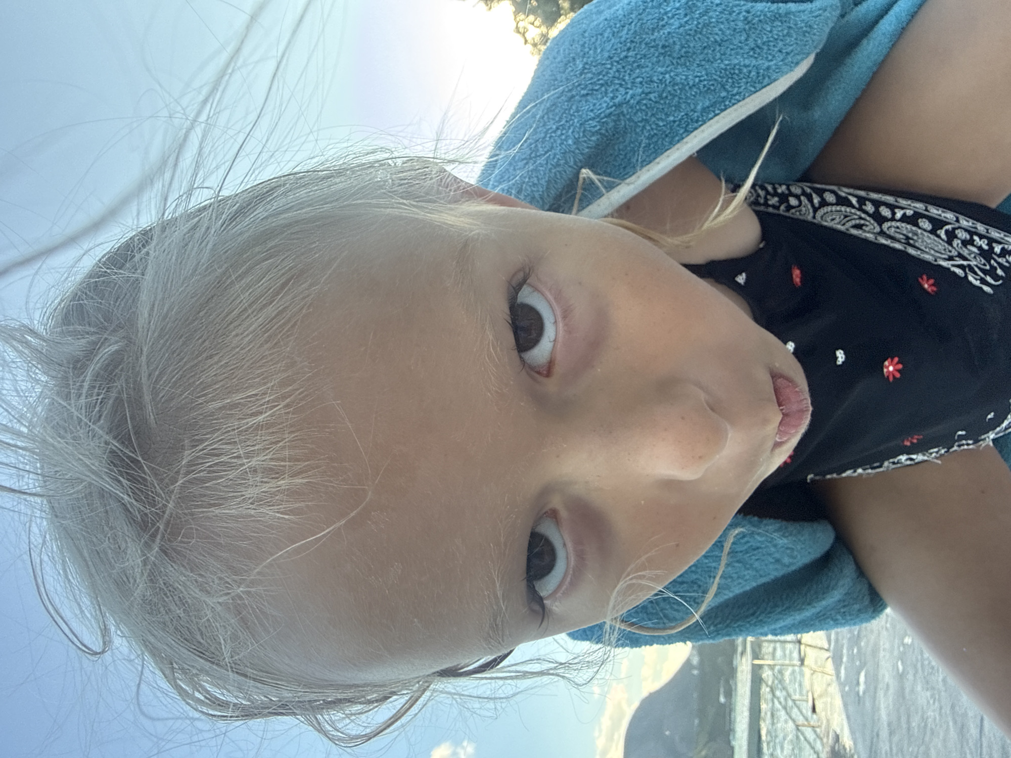 A close-up, vertically oriented photograph shows the face of a young child. The child has fair skin, light blonde hair that appears windblown, and a light-colored eye with dark pupils. The child is wearing a black top with a decorative pattern along the neckline, which includes small red and white floral designs. A blue towel is draped around the child's shoulders. The background is out of focus but appears to be an outdoor setting with a bright sky and some distant landscape elements, possibly mountains. The lighting suggests it is daytime, with the sun creating a bright, slightly hazy atmosphere. The child's expression is a direct gaze towards the camera, with their mouth slightly open. The overall impression is a candid moment captured outdoors.