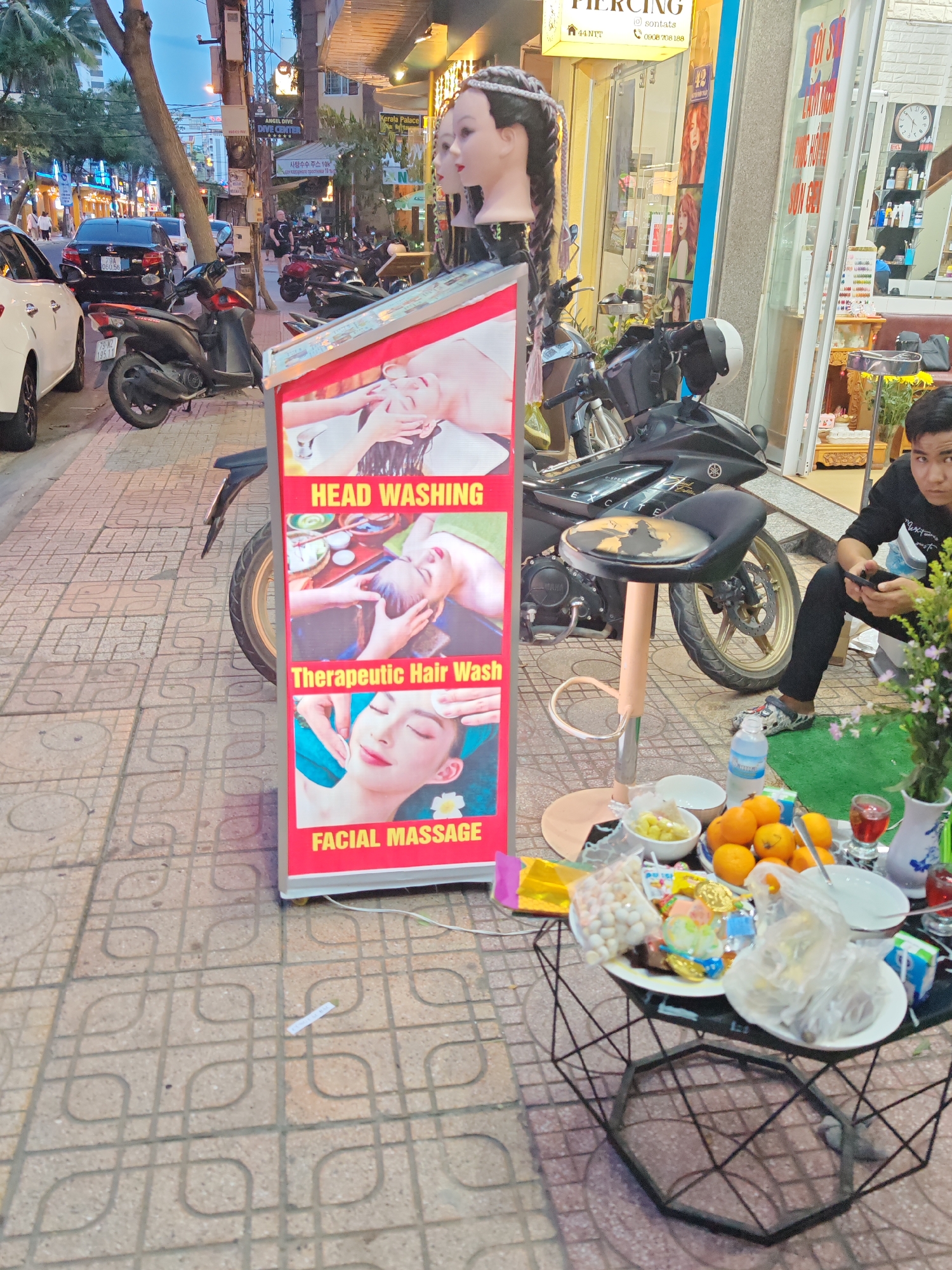 The image depicts a street scene in Nha Trang Ward, Vietnam, focusing on a salon's advertising display. In the foreground, a tall, red advertising stand features three panels with images of women receiving spa treatments. The top panel reads "HEAD WASHING," the middle panel reads "Therapeutic Hair Wash," and the bottom panel reads "FACIAL MASSAGE." To the right of the stand, a black motorcycle is parked, and a small table laden with food, including oranges, bowls of fruit, and wrapped snacks, is visible. A person is seated on the ground to the right, looking at a phone. In the background, other motorcycles are parked along the sidewalk, and shopfronts with signage are visible, indicating a commercial area. The pavement is paved with square tiles.