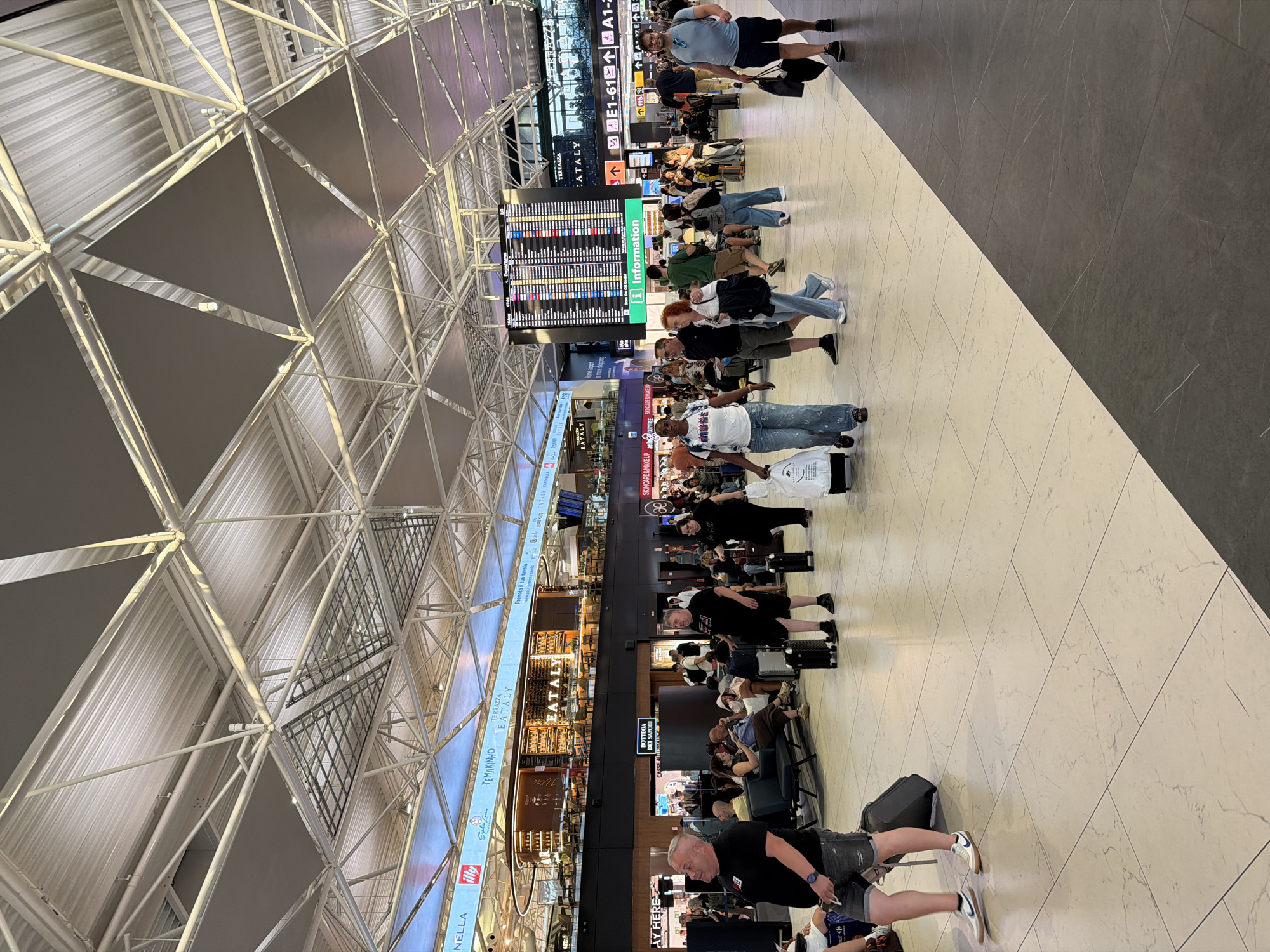 The image depicts a bustling interior scene within an airport terminal in Fiumicino, Italy. The architecture features a high, angular ceiling with exposed metal trusses and numerous triangular panels. Natural and artificial light illuminates the spacious concourse, where passengers are seen walking, standing, and sitting.
In the foreground, a man in a black t-shirt and denim shorts is visible from the waist down, pulling a grey rolling suitcase. To his right, another man in a black t-shirt and shorts also has a black rolling suitcase. Further to the right, a woman in a black t-shirt and black pants is pulling a white rolling suitcase.
Midground, a large departure board displays flight information, with "Departures" clearly visible at the top. Several shops and restaurants are visible on the upper level, including one with a prominent "EATALY" sign. People of various ages and ethnicities are dispersed throughout the terminal, some with luggage, suggesting travel. The floor is tiled with large, light-colored square tiles, with a dark gray tiled area diagonally bisecting the lower portion of the image. The overall atmosphere is one of transit and activity typical of an international airport.