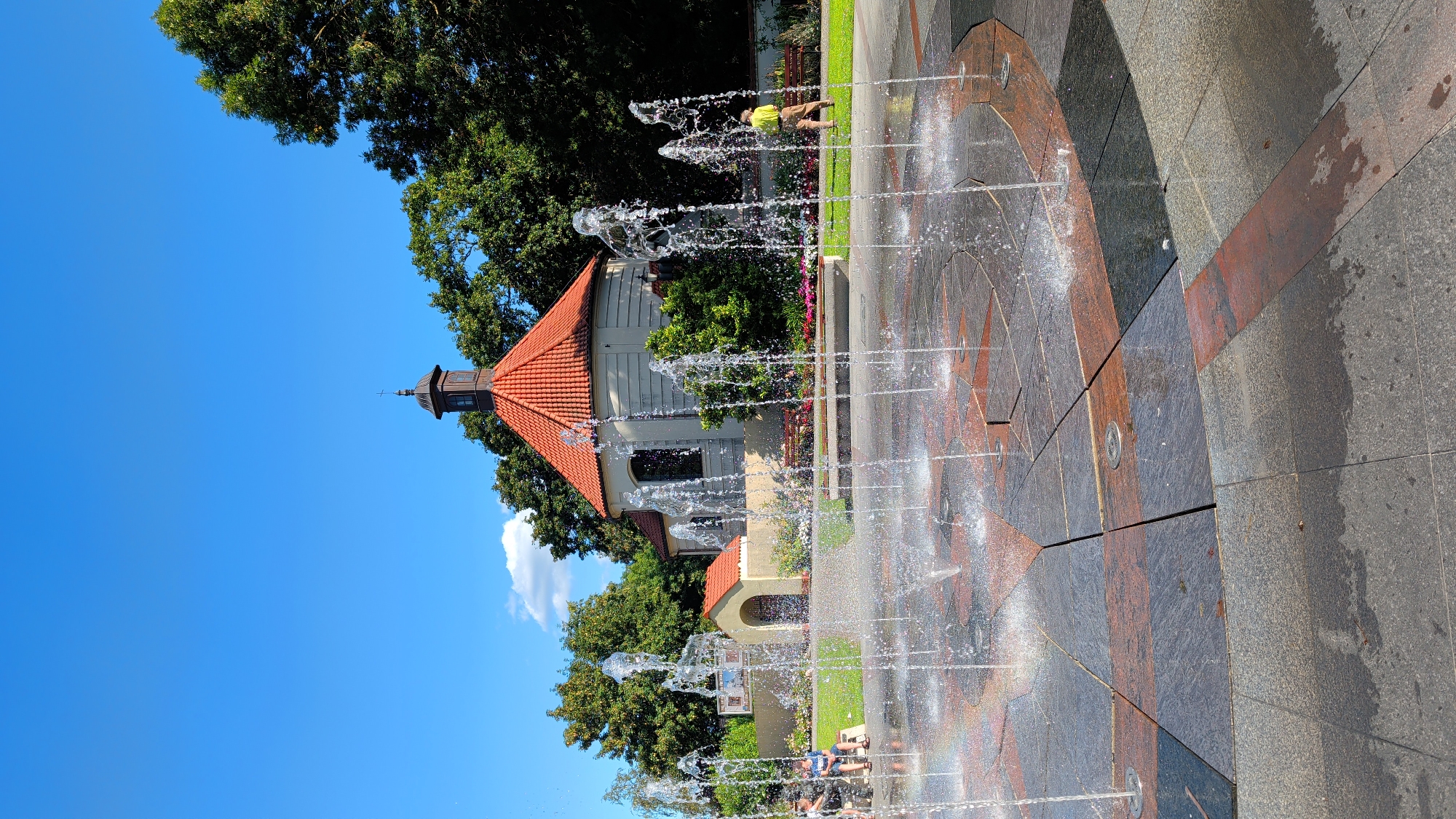 The image depicts a public fountain located in Pułtusk, Poland, likely in a park or plaza area. In the foreground, a modern fountain sprays water upwards, creating a dynamic water display. The fountain's base is paved with a mosaic of granite tiles in various shades of gray and reddish-brown, forming a curved pattern.
In the midground, a man in a light green shirt and brown pants stands facing away from the camera, near a bench. Behind him, a picturesque building with a red tiled roof, possibly a chapel or a small church, is visible. The building has a distinctive tower topped with a cross. Lush green trees and flower beds with colorful blooms surround the building and extend into the background, suggesting a well-maintained outdoor space.
On the left side of the midground, two individuals are seated on a bench, partially obscured by the fountain's water spray. The sky above is a clear, vibrant blue with a few scattered white clouds. The overall scene conveys a sense of a pleasant, sunny day in a public recreational area.