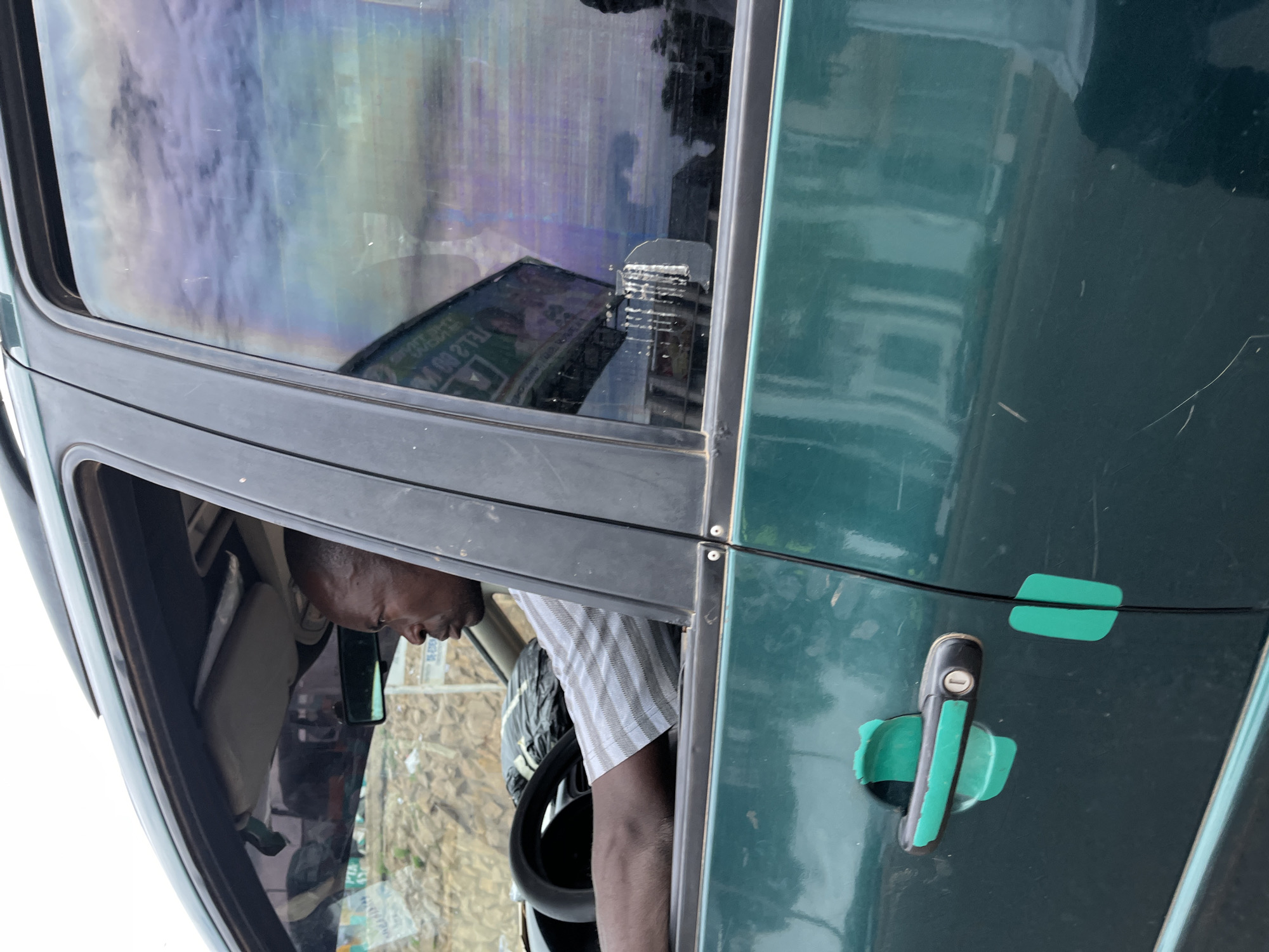 An adult male with dark skin and short hair is depicted in the driver's seat of a dark green vehicle, consistent with an SUV or similar model. He is viewed in profile, looking towards the right of the frame, with his right hand on the steering wheel. He wears a light-colored, short-sleeved shirt with vertical stripes. Inside the vehicle, a black plastic bag rests on what appears to be the front passenger seat or dashboard. The sun visor is in the upright position, and a rearview mirror is visible.
The exterior of the vehicle's front driver's door features a black door handle with a visible keyhole, partially covered or embellished with teal-colored tape or material. Additional teal tape pieces are applied to the door panel near the door seam. Scratches are visible on the lower section of the dark green door. The side mirror on the driver's side is absent or severely damaged, with adhesive residue and fragments visible on the lower rear portion of the window frame.
The driver's side rear passenger window displays significant reflections, including a cloudy, light-colored sky with hues of purple and blue. A large, inverted reflection of a billboard with dark green and white elements is prominent, displaying partial text such as "TEL 2 00". Other reflections suggest urban structures and ambient light.
Through the front windshield, a background of rocky or hilly terrain, possibly an embankment or construction site, is visible, along with a white signpost. The scene is located in the city of Jos, Nigeria.