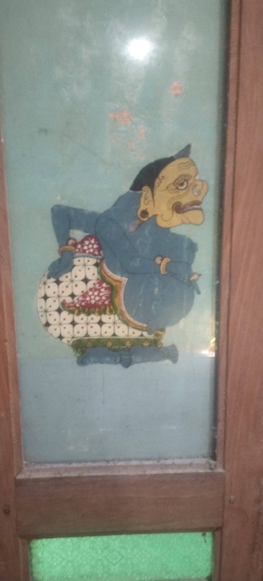The image shows a painting of a stylized character, possibly from traditional Indonesian Wayang Kulit puppetry, on a frosted glass pane framed by wooden borders. The character is depicted with a large, rounded body, blue skin, and a yellow face with prominent features. It wears a patterned garment that resembles batik and has its arms crossed. The background of the painting is a pale blue. The frosted glass pane is part of what appears to be a door or window in Daleman, Indonesia. The wooden frame is weathered and the lower section of the frame contains a pane of textured green glass or material. A bright light source is reflecting off the glass, obscuring some details in the upper portion of the painting.