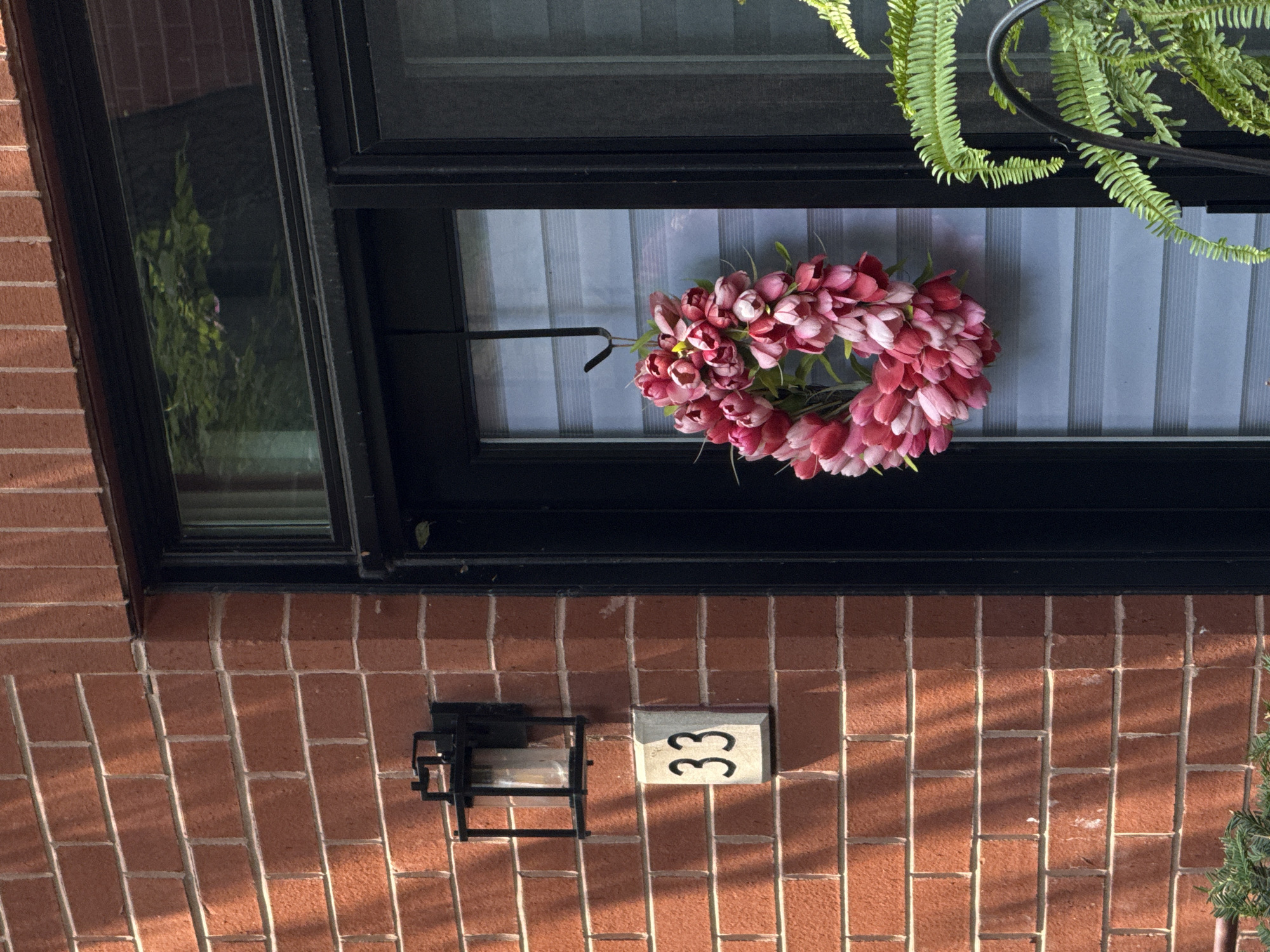 A tulip wreath hangs on a door next to a brick wall with the house number 33. A black outdoor light fixture is mounted on the brick wall above the house number. A fern plant is visible in the foreground on the right side of the image. The scene is set in Chicago, United States of America.
