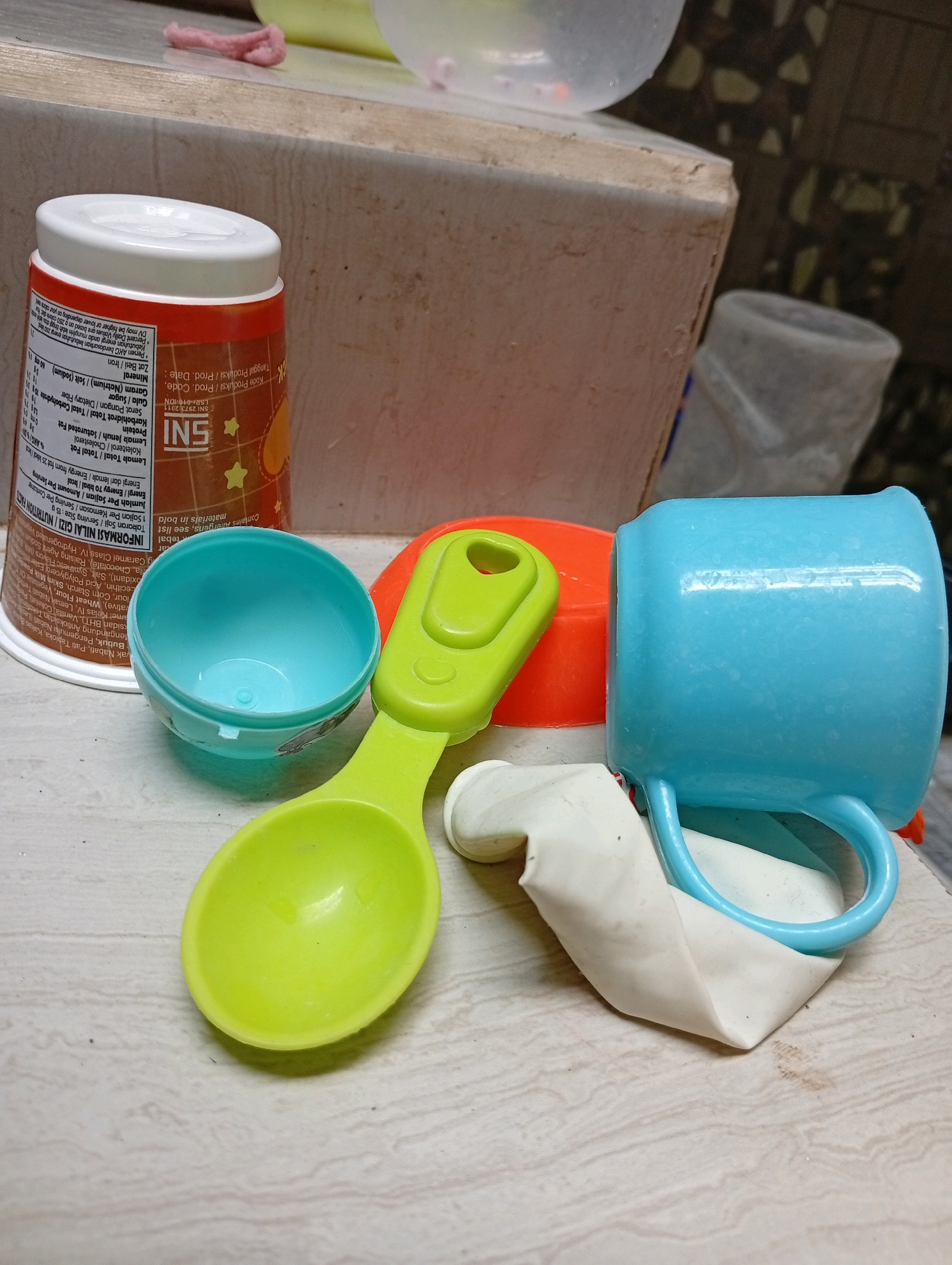 The image shows a collection of colorful children's toys and a container arranged on a textured surface. In the foreground, a bright green plastic scoop is positioned horizontally, with its bowl facing upwards and slightly to the left. Behind the scoop, a teal-colored plastic cup with a handle is tilted to the right, resting on a deflated white balloon. An orange plastic lid is visible behind the scoop and cup. To the left, a cylindrical container with a white lid and orange accents is partially visible, featuring text and graphics. A smaller teal bowl is placed to the left of the green scoop, its opening facing upwards. The background reveals a slightly out-of-focus view of a textured surface, possibly a wall or a piece of furniture, with subtle patterns. The overall scene appears to be an indoor setting, with the toys likely belonging to a child. The provided location, Kebondalem, Indonesia, suggests this is a domestic setting within that city.