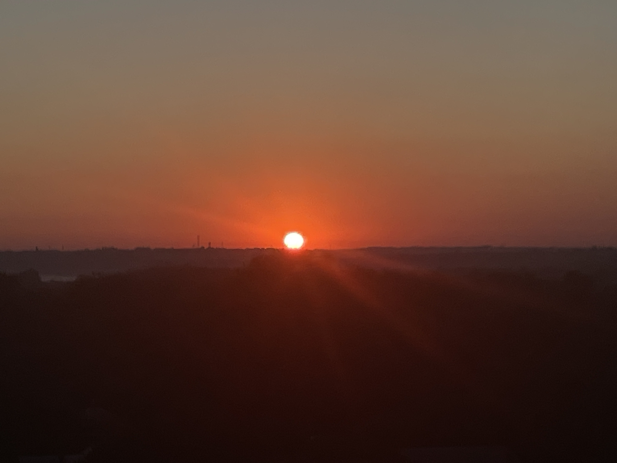This image captures a sunset over Novocherkassk, Russia, viewed from a high vantage point. The sun, a bright orange disc, is setting on the horizon, casting a warm glow across the sky. The sky transitions from a pale yellow near the horizon to a muted gray at the top. In the distance, a faint line of trees and buildings are silhouetted against the orange sky, suggesting the presence of the city. The foreground is dominated by a dark, shadowy landscape, likely hills or forests, with the light of the setting sun creating rays that emanate from behind the sun. No people or specific objects are clearly discernible due to the low light conditions and the distance. The overall impression is one of a serene and atmospheric sunset over a semi-urban landscape.