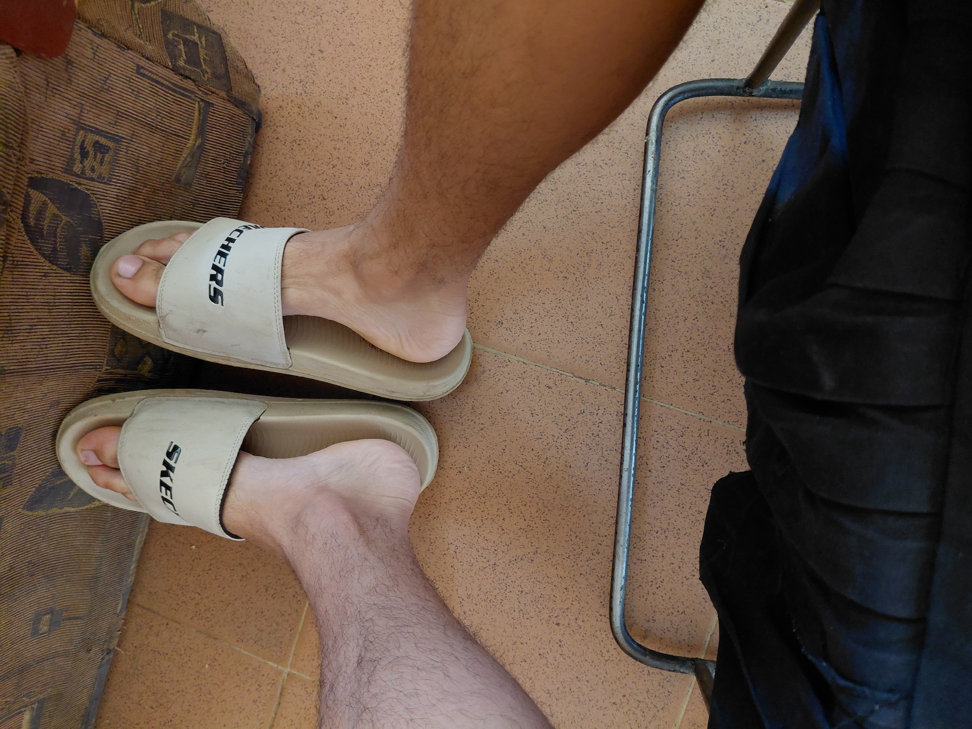 A top-down view captures two pairs of bare feet wearing identical light-beige Skechers slide sandals. The left leg and foot exhibit a lighter skin tone with sparse hair, while the right leg and foot are significantly darker with denser hair. Both pairs of feet are resting on a light brown tiled floor featuring subtle speckles and visible grout lines. Behind the feet, an upholstered surface with vertical ribbing and abstract dark patterns is visible, indicating furniture such as a couch or chair. In the foreground, a curved, silver-colored metal bar, likely part of a chair frame, extends horizontally across the lower portion of the image, beneath which dark fabric, possibly clothing, is present. The scene is located in Unknown, Unknown.