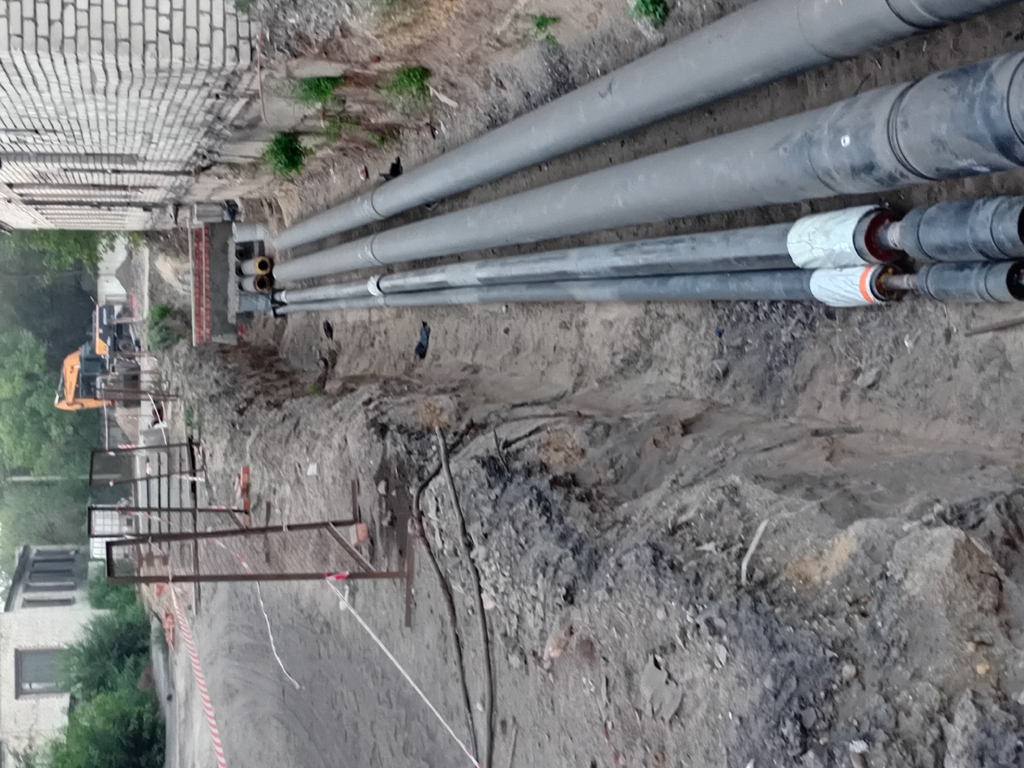 The image depicts an outdoor construction site with a trench being dug alongside a brick building. The trench contains several large, grey pipes, likely for utility services. There are also smaller, black pipes with orange and white insulation visible. In the background, an excavator is visible, suggesting ongoing construction work. A red and white striped caution tape is strung along a temporary fence, demarcating a work zone. The ground is composed of dirt and gravel. There are no people or animals visible in the scene. The environment appears to be a semi-urban or industrial area. The presence of construction equipment and the trench indicates that infrastructure work is underway in Semey, Kazakhstan.