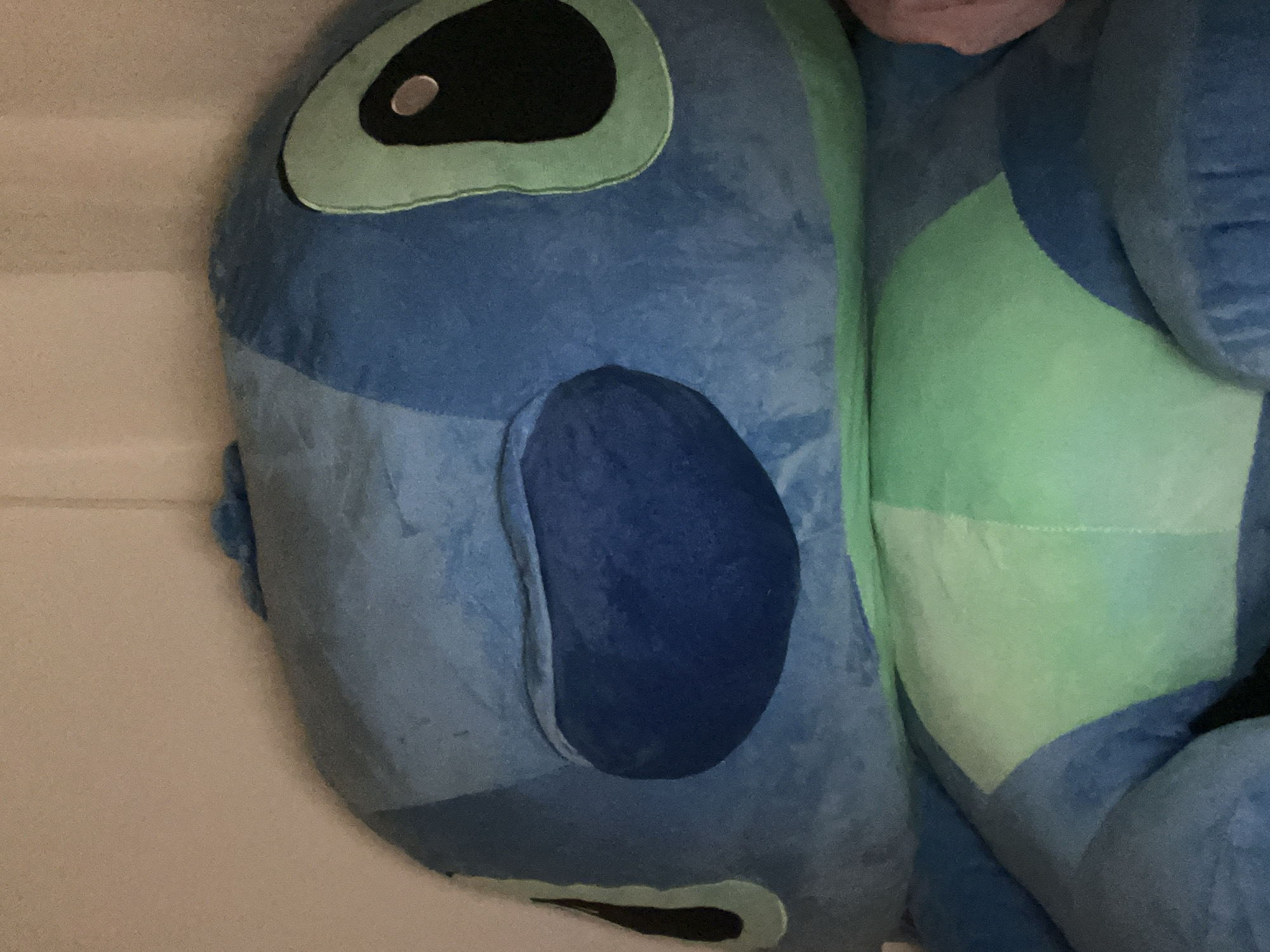 This close-up image features a large plush toy, strongly resembling Disney's Stitch, resting in an indoor setting. The toy is predominantly blue, with varying shades from a medium blue main body to darker blue patches on its ear and face, and light green accents.
Key features of the plush toy include a prominent black oval eye with a reflective white or silver pupil, surrounded by a light green outline. Below the eye, a dark blue, rounded ear is visible. Another dark blue patch is seen on what would be the nose area. The body of the toy displays segments of light green fabric stitched into the blue material, suggesting the character's distinct pattern. The material appears soft and fuzzy, indicative of a plush toy.
The background is a plain, light, off-white surface, possibly a wall or bedding, with faint horizontal lines. The image is oriented with the top of the toy's head towards the left. A small, light-skinned portion of a person, likely an arm or hand, is visible at the very bottom right of the frame, suggesting the presence of an individual near the toy. No specific activity is taking place; the toy appears to be static and at rest. There is no visible text in the image. The provided context indicates the location is Plovdiv, Bulgaria.