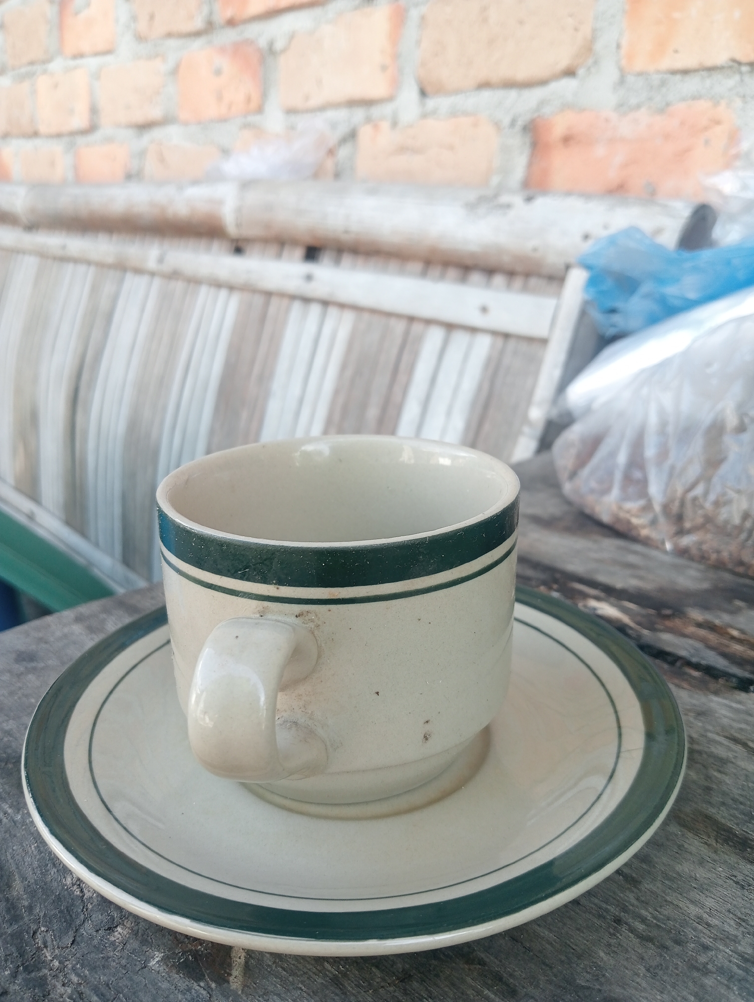 A close-up, eye-level shot captures a light beige ceramic mug with two dark green bands near the rim, resting on a matching saucer. The mug has a pale beige handle. The setting appears to be outdoors, on a weathered wooden table. In the background, a brick wall with varying shades of orange and grey bricks is visible, above a slatted wooden bench. To the right, a clear plastic bag filled with small, brown, granular items is partially visible. The overall lighting suggests it is daytime. The location is identified as Kota Negara Timur, Indonesia.