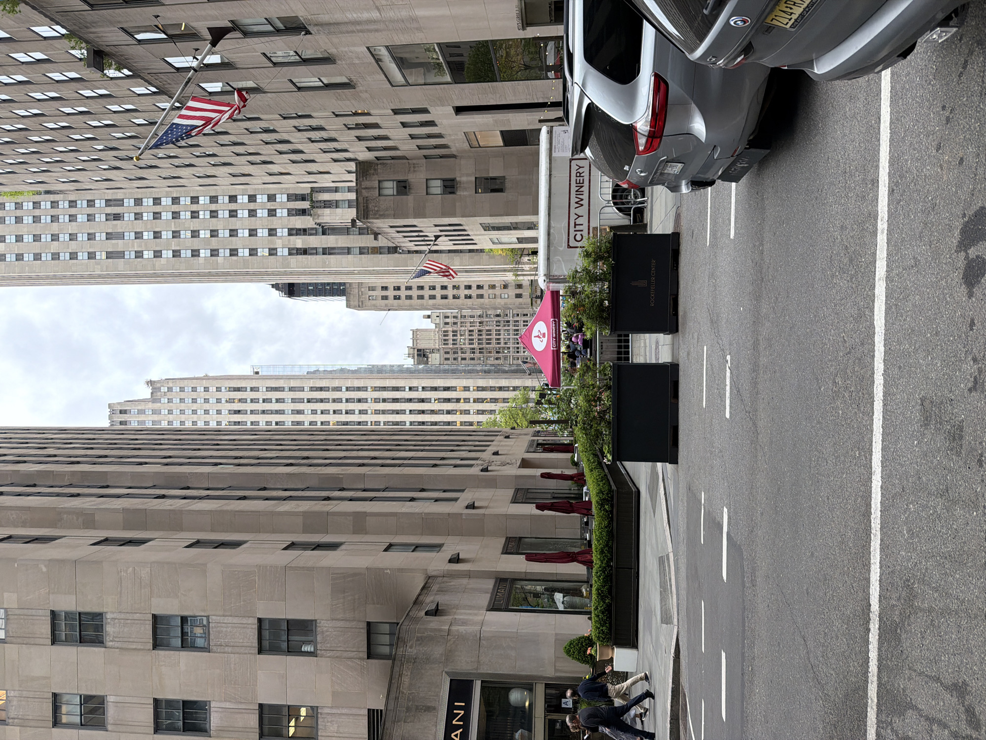 This media file captures a vertical urban street scene, likely in New York City near Rockefeller Center. The image is originally rotated 90 degrees counter-clockwise.
In the foreground, an asphalt street with white parking lines runs horizontally. Two silver cars are parked on the right side of the street, including an SUV whose license plate holder reads "THE ART OF PUTTY" and a partial license plate shows "Z14 R". Adjacent to the street, a sidewalk features black planter boxes filled with greenery, one clearly labeled "ROCKEFELLER CENTER". Further along the sidewalk, an outdoor dining area is set up with red umbrellas and a prominent pink canopy or tent. Text on the tent and a nearby trailer indicates "CITY WINERY," and "US GRILLE" is visible on a menu or sign within the tent. Several people are seen walking on this sidewalk.
Tall, multi-story buildings with numerous windows and stone or concrete facades dominate both sides of the street and extend into the background. One building on the left shows the partial word "ANI", suggesting "LIMANI" restaurant. Two American flags are displayed from poles on different buildings, adding to the urban landscape. The sky is overcast, indicating a cloudy daytime setting.