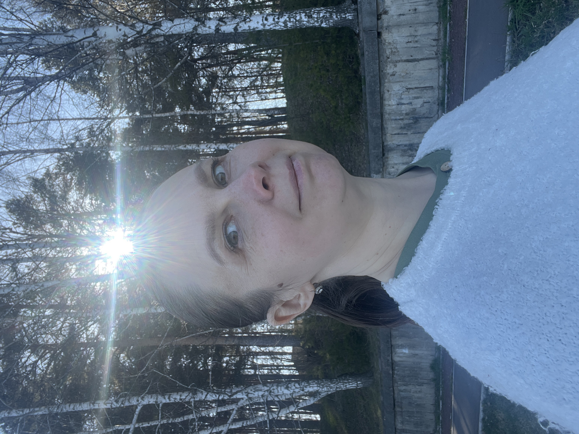 A young woman is pictured outdoors. She is wearing a light-colored, possibly white or light blue, textured sweater or jacket over a green garment, which may be a shirt or dress. Her hair is pulled back, revealing an earring. She is facing forward with a slight tilt to her head. The background features birch trees and what appears to be a concrete fence or barrier. The sun is visible behind the trees, creating a lens flare. The location is likely in the city of Chaikovsky, Russia.
