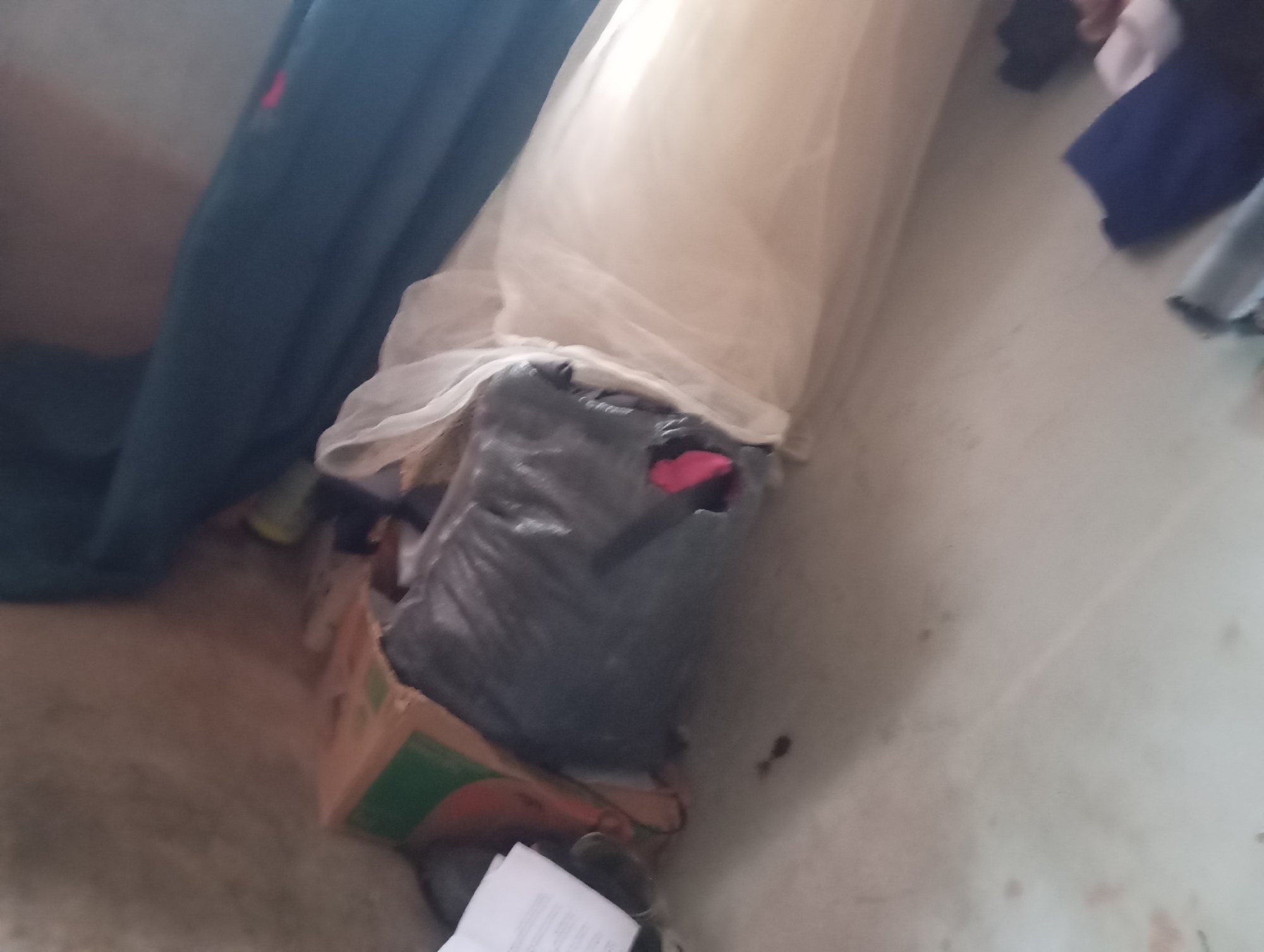 The image depicts a dimly lit indoor scene, possibly a storage area or room, with the floor appearing to be concrete. Dominating the center is a large, black plastic-wrapped bundle, lying horizontally on top of an open cardboard box. The cardboard box is partially visible, showing some green and orange markings. To the left of the bundle is a dark blue fabric, possibly a curtain or drape, which extends vertically. Partially obscuring the bundle is a sheer, light-colored fabric, likely a veil or sheer material, draped over it. A bright pink piece of fabric is visible protruding from the top of the black bundle. The background is indistinct, but suggests further clutter or other draped fabrics. There are no people clearly visible in the image. The overall impression is one of disarray and storage. The context of Jalingo, Nigeria, suggests this scene is located within that city.