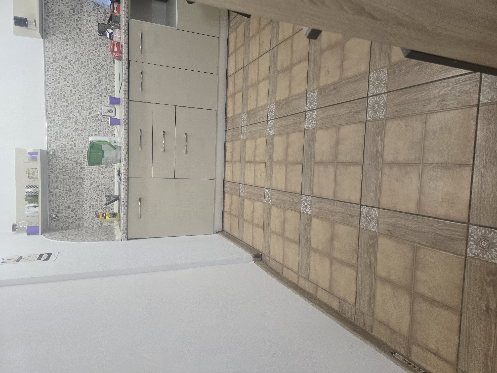 The image depicts a kitchen area. The flooring consists of square tiles in a tan color with a wood-grain pattern bordering some of the squares. The walls are white, and a white baseboard runs along the floor. In the background, there is a countertop with a mosaic tile backsplash. On the countertop, various items are visible, including a green water filter pitcher, two purple cups, a white appliance, and some packaged goods. Below the countertop are light-colored cabinets with silver handles. A sink with a faucet is also present. The overall impression is that of a functional, albeit somewhat sparsely decorated, kitchen. The image does not provide any specific geographical indicators that would allow for its localization in Ulan-Ude, Russia, or any other city.