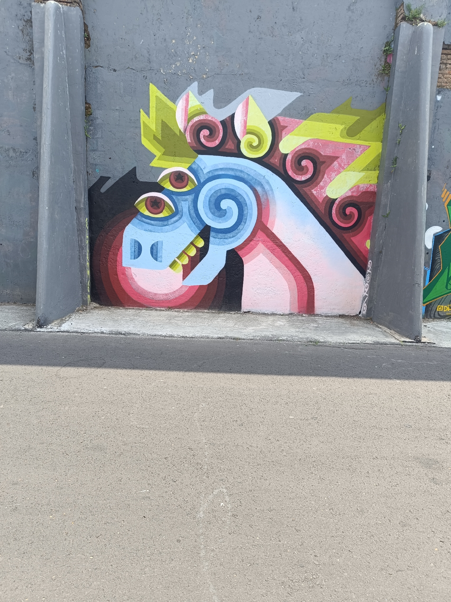 This image depicts a brightly colored graffiti mural on a concrete wall, situated in the city of Yogyakarta, Indonesia. The mural features a stylized, abstract depiction of a horse's head, characterized by bold lines and vibrant colors. Dominant colors include electric blue, pink, yellow, and red, with swirling patterns and geometric shapes. The artwork is framed by two large, grey, pillar-like structures that extend vertically along the sides of the wall, suggesting the mural is part of a larger urban landscape, possibly an underpass or a designated street art area. In the foreground, a section of asphalt road is visible, casting a distinct shadow from an unseen object. The ground where the mural meets the road is paved with concrete. The overall scene captures a piece of contemporary urban art within the context of Yogyakarta's street culture.
