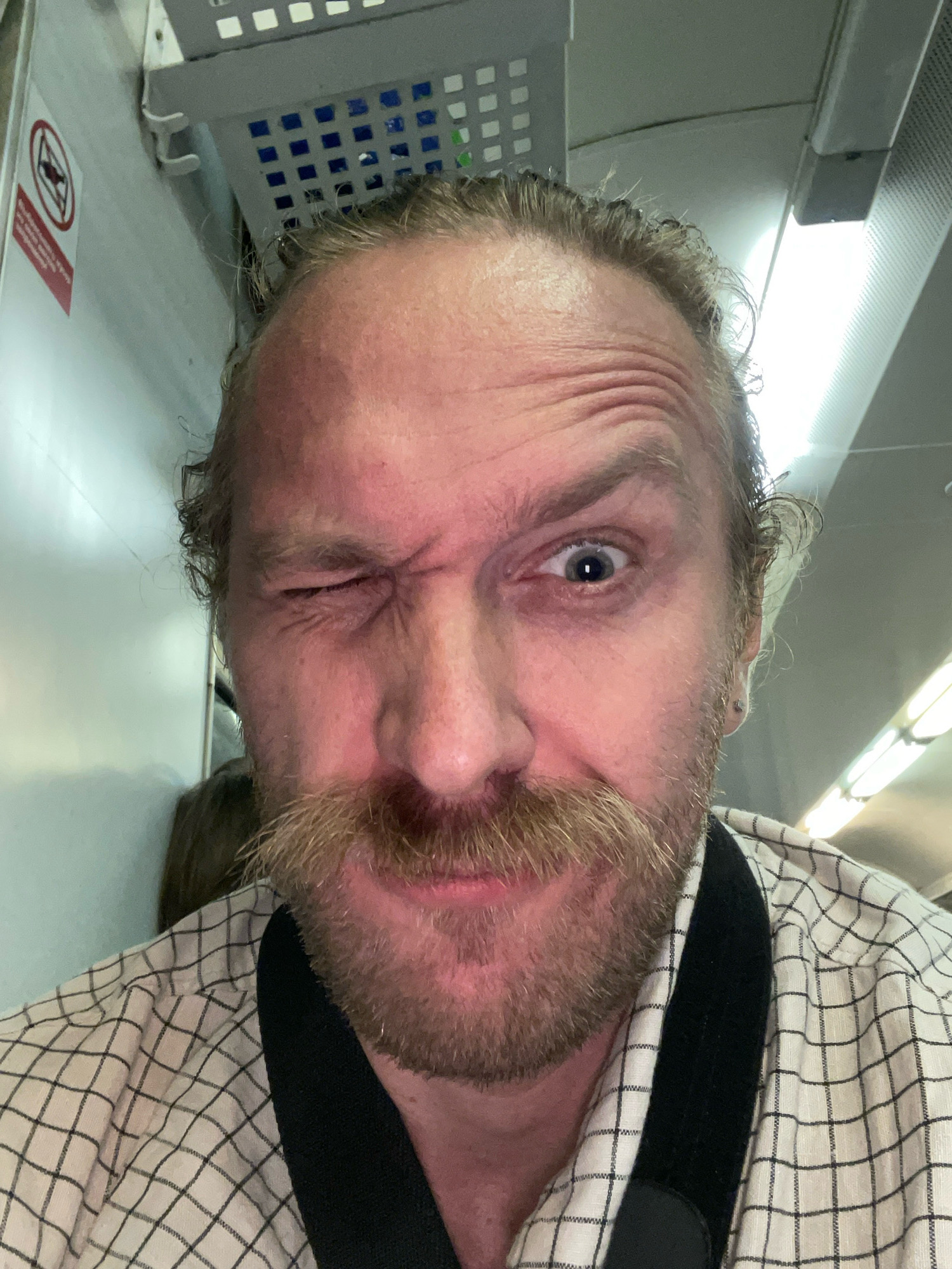 A man is taking a selfie in what appears to be a train carriage. The man has blonde hair pulled back in a bun, a prominent mustache, and stubble. He is winking with his left eye and his right eye is open. He is wearing a white shirt with a black grid pattern. The environment appears to be inside a train, with overhead luggage racks and fluorescent lighting visible. There is a "no smoking" sign on the wall to the left of the man. The image does not provide enough information to determine the specific location in the city of Kornevo, Russia.