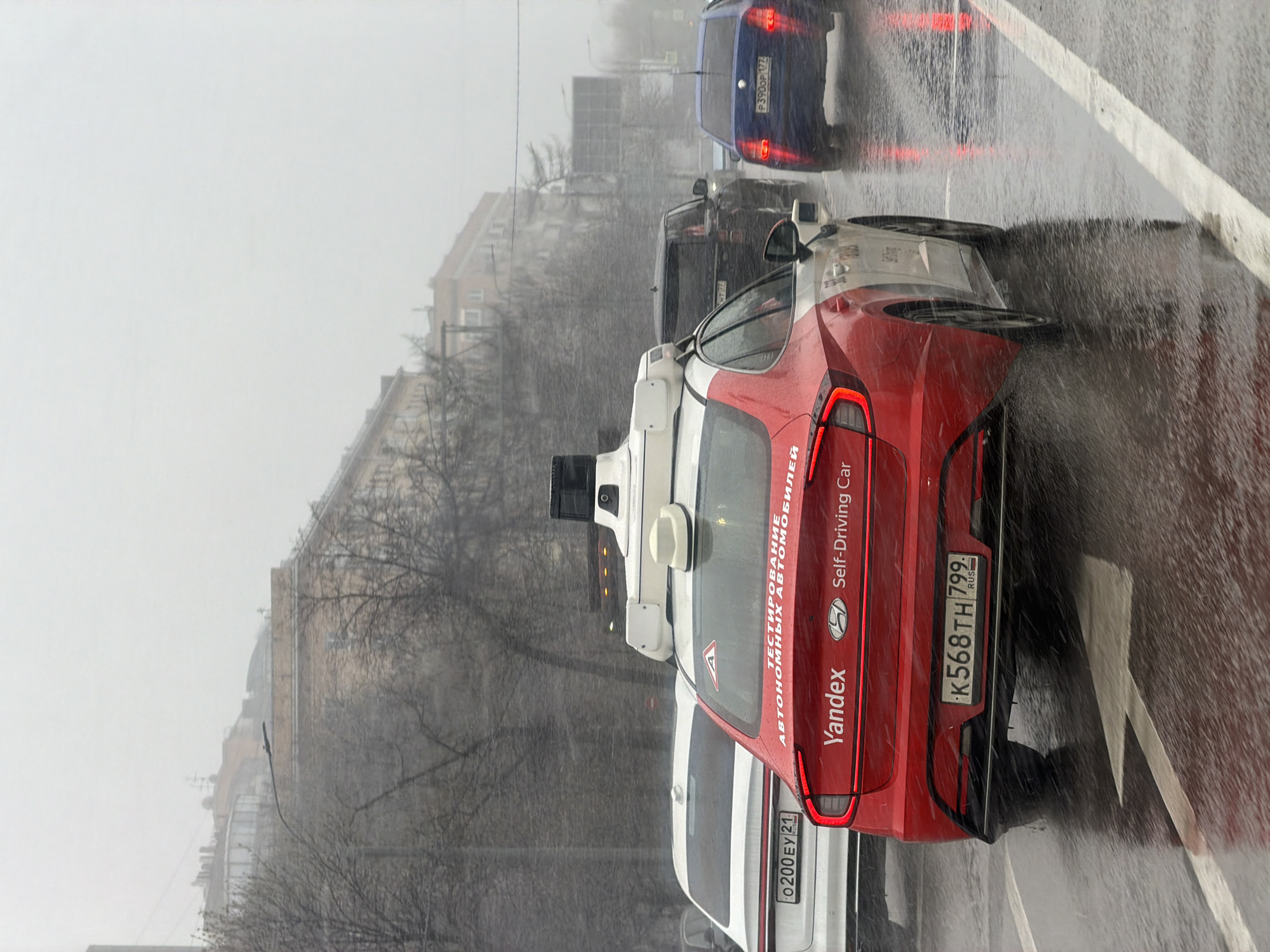 The image shows a red Yandex self-driving car in Moscow, Russia, amidst heavy rain. The car is stationary on a street marked with white lines, part of a traffic queue that includes a blue car and a black car. The backdrop features buildings and bare trees obscured by the rain and fog. The number plates on the Yandex car read "K568TH 799" and "O200EY 21," with the inscriptions "Yandex Self-Driving Car" and "АВТОНОМНЫХ ОВАНМОБИЛЕЙ" visible on its body. The blue car ahead shows the license plate "P3900P 177". The environment suggests a busy urban setting under adverse weather conditions.