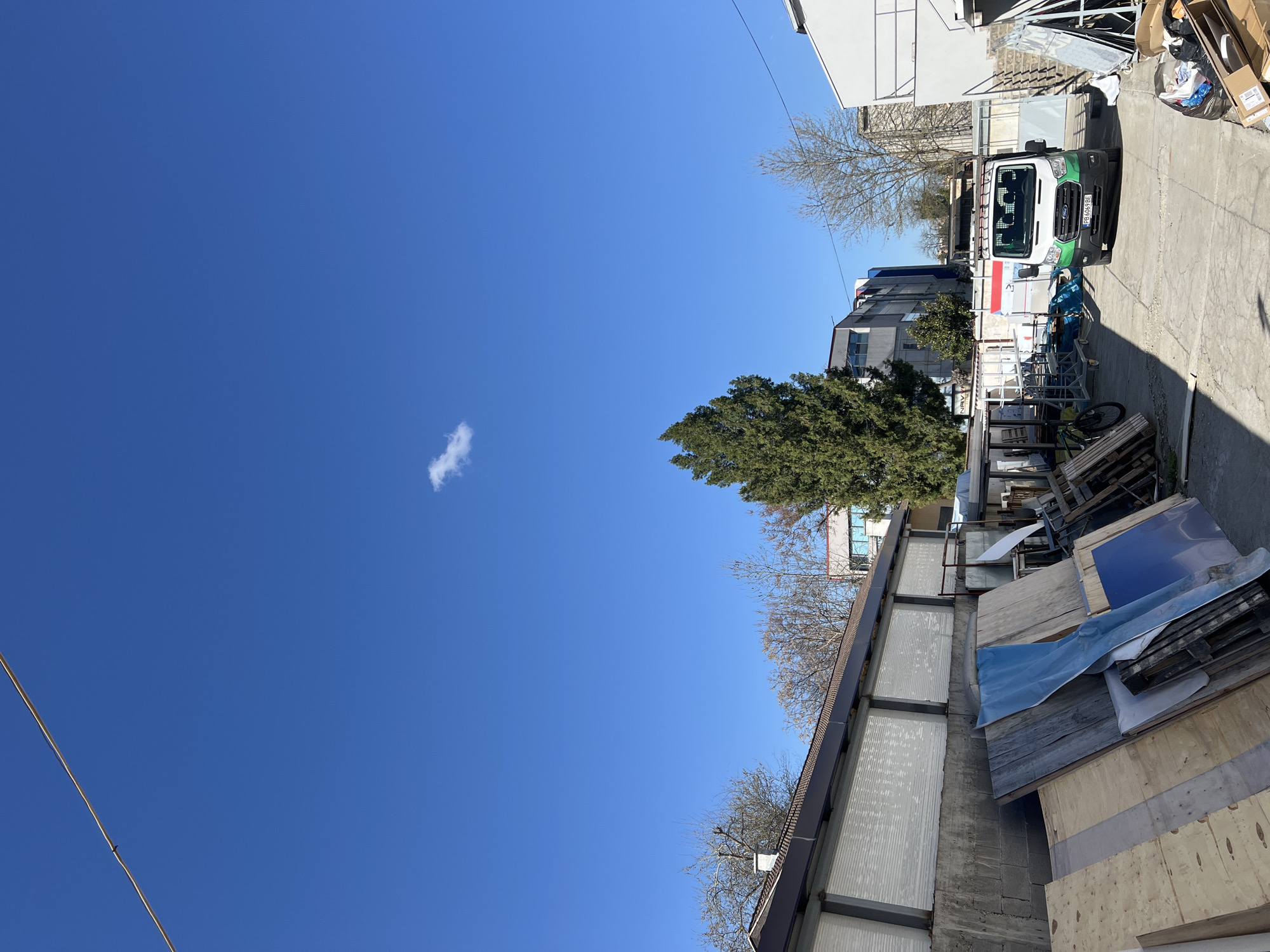 The image shows an outdoor scene, likely in an industrial or semi-industrial area of Plovdiv, Bulgaria. A bright blue sky with a small cloud occupies the upper portion of the image. Buildings of varying styles are visible. A white Ford Transit van is parked along the right side of the image. A cluttered area with pallets, construction materials, and debris is visible to the lower right of the van. The foreground includes corrugated and wooden materials, possibly part of a storage structure or construction site. A bare tree is visible near the building. A wire or cable is seen in the upper left portion of the image. No people are visible.