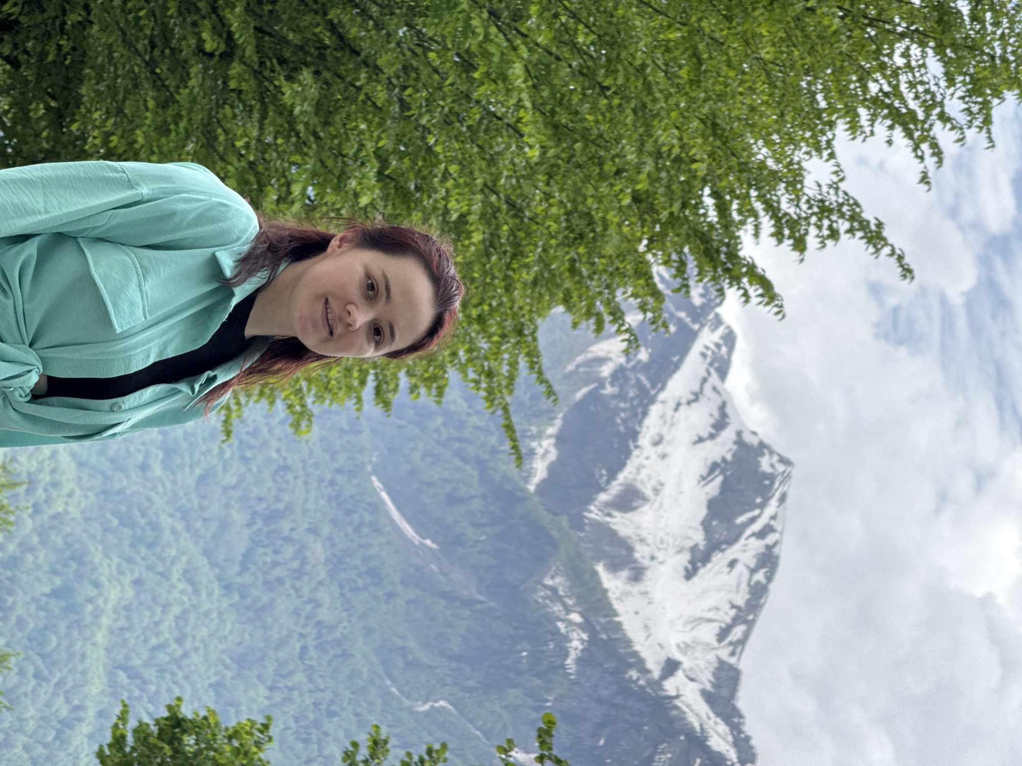 The image shows a woman standing against a backdrop of mountains in Krasnaya Polyana, Russia. The woman has reddish-brown hair and is wearing a light green shirt over a black top. She is smiling slightly and looking towards the viewer. Behind her, there is a lush green mountain range and a taller, snow-capped mountain. There are trees in the foreground partially obscuring the view, and some clouds in the sky.