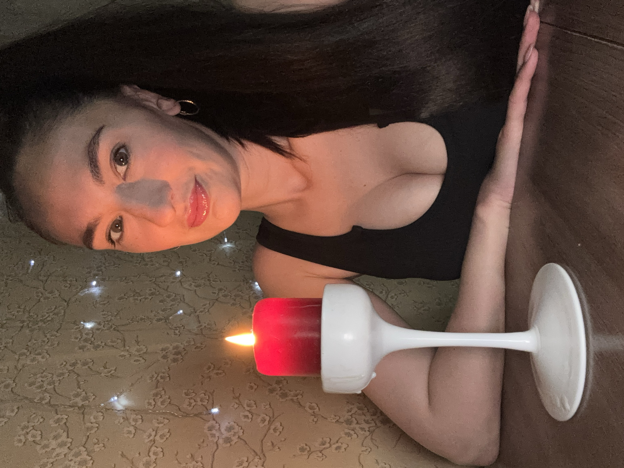 An indoor, low-light photograph, likely taken in Sochi, Russia, captures a woman reclining with a lit red candle. The woman, with long dark hair, is positioned on her side, looking towards the viewer with a gentle, serene expression. She wears a dark top, possibly a camisole, and a gold hoop earring is visible on her left ear. Her lips are a glossy pink, and her eyes are open.
Her right arm is bent, and her left arm extends forward, resting on a dark, smooth surface, possibly a wooden table or floor. The red pillar candle, actively burning, sits in a white, goblet-shaped candle holder, casting a warm glow that illuminates parts of her face and the immediate surroundings.
She is lying on a patterned fabric surface, possibly a rug or blanket, which features a subtle floral or leaf design. Scattered across this surface are small, white LED string lights, creating soft, sparkling points of light and bokeh effects, contributing to a cozy and intimate atmosphere. The overall scene suggests a relaxed evening or night setting. There is no visible text in the image.