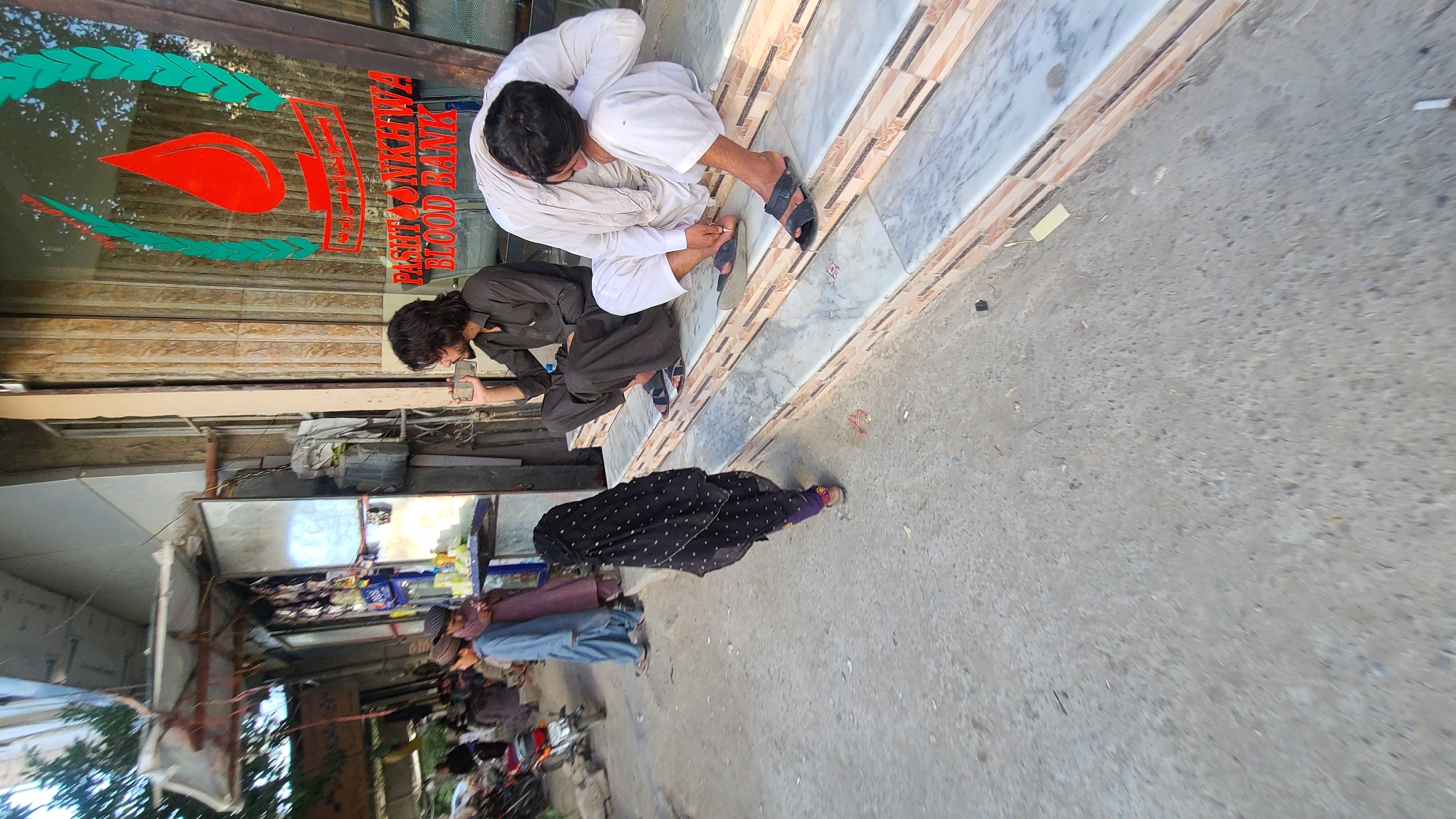 Outside the Pashtoonkhwa Blood Bank in Quetta City Tehsil, Pakistan, two men are crouched down, with one appearing to use a mobile phone. A woman in a black dotted veil stands nearby. Further down the street, there are other individuals and parked motorbikes. The environment is a busy urban street with buildings, shops, and street debris.