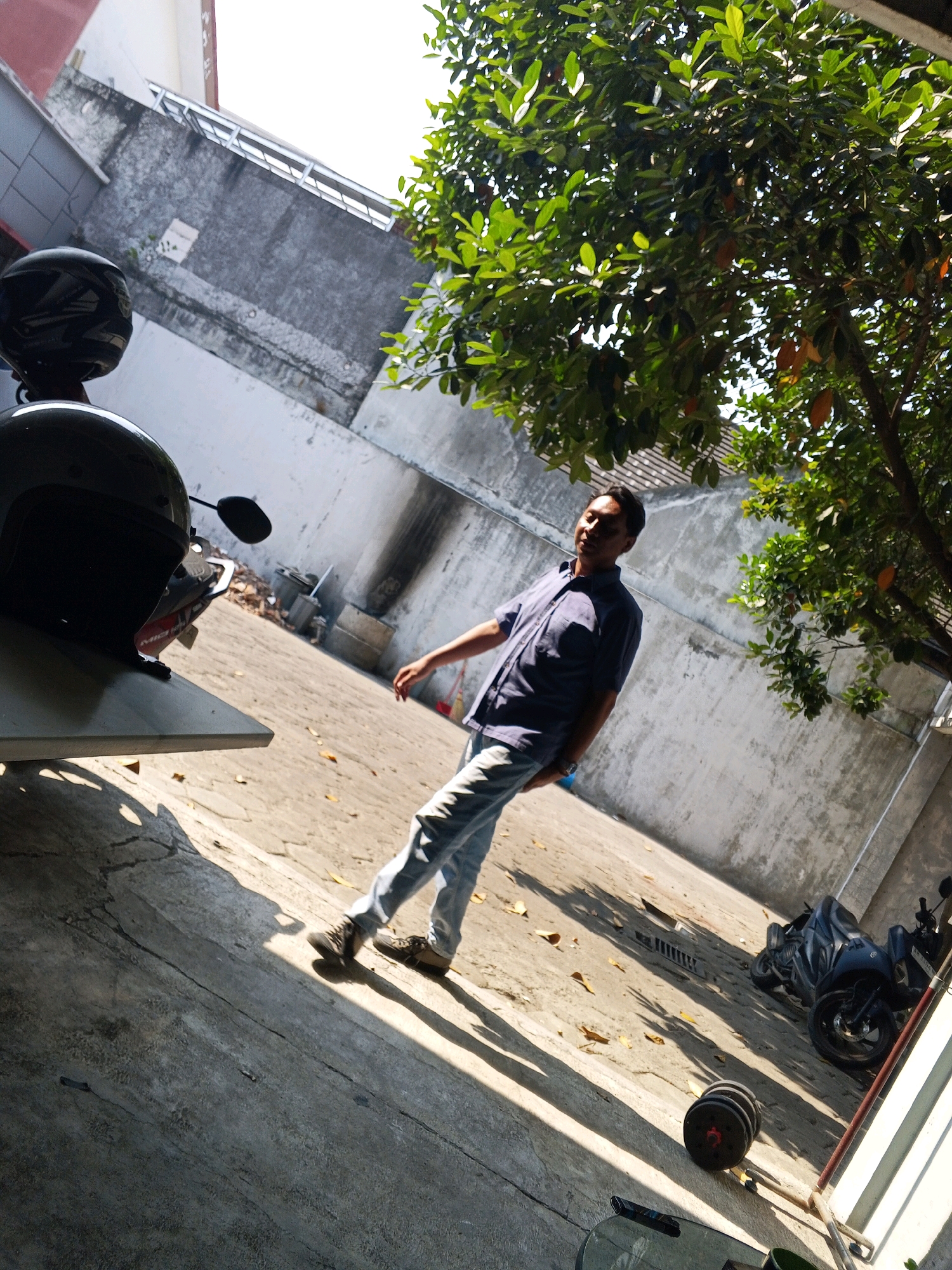 A man stands in a concrete courtyard in Condongcatur, Indonesia, bathed in sunlight filtering through a large tree with green leaves and some visible fruit. He is wearing a dark blue collared shirt and light blue jeans, with dark shoes. To his left, a black motorcycle helmet rests on a table. In the background, a gray concrete wall runs along the side of a building, with a second motorcycle parked further down. The ground is a mixture of concrete and what appears to be paving stones, with fallen leaves scattered across it. The scene is characterized by strong shadows cast by the tree and the man.