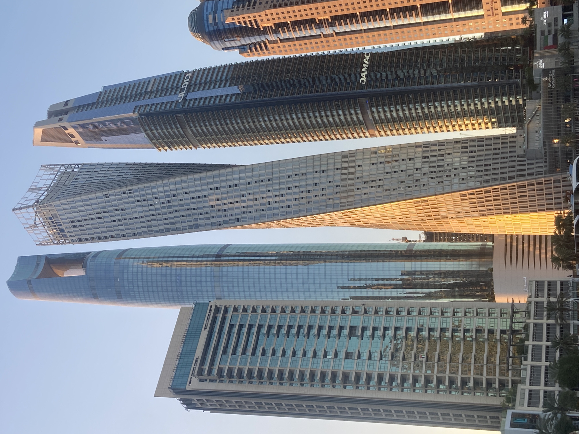 The image shows a low-angle view of several skyscrapers in Dubai, United Arab Emirates, against a clear sky. The buildings feature modern architectural designs, with glass facades and varying heights. One notable building has a distinct twisting shape. The colors in the image range from cool blues of the glass to warm hues reflecting the setting sun. The "DAMAC" logo is visible on one of the buildings. Other signs in Arabic and English are visible along the base of the buildings. Palm trees and other vegetation are also visible along the base.