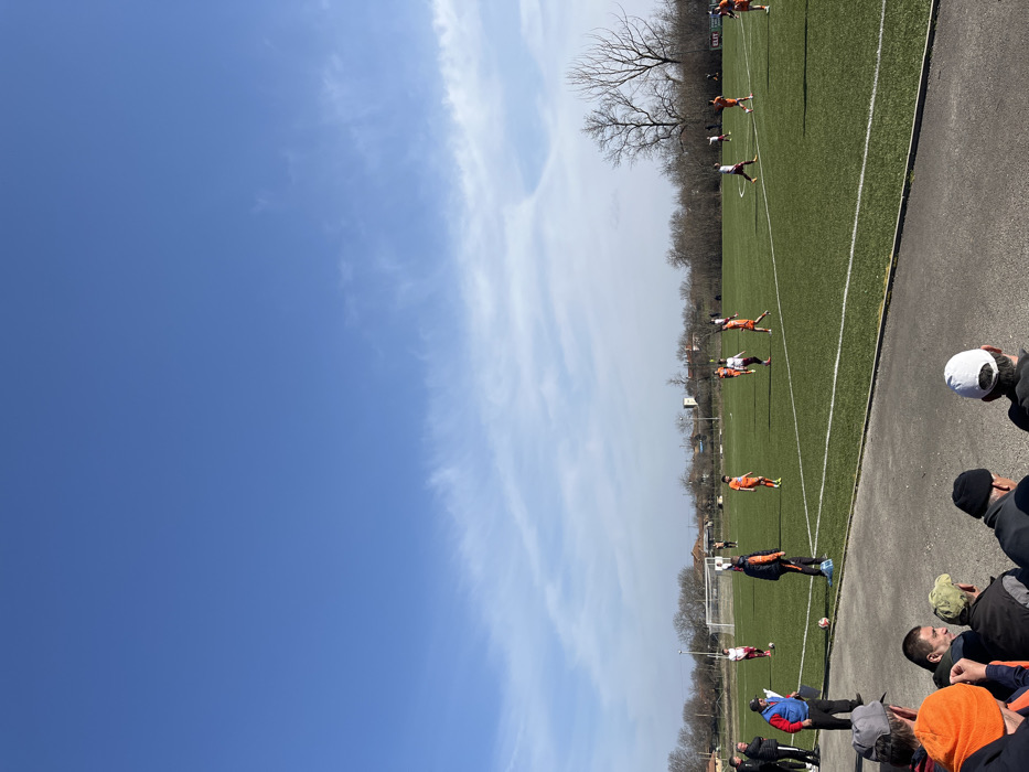 This image depicts an outdoor football match in progress on an artificial green turf pitch located in Kliment, Bulgaria. Approximately 10-12 players are visible, divided into two teams: one wearing orange kits and another in white shirts with maroon shorts or accents. Players are distributed across the field, with white lines marking the pitch boundaries.
In the foreground, a group of male spectators, some wearing hats, are seated or standing, observing the game from behind a concrete barrier. Near the sideline, a male individual in a blue vest and red shirt stands, appearing to be a coach or official, observing the play. A football rests on the pitch near the sidelines in the mid-ground.
The background features distant residential buildings, including a structure resembling a church tower, and numerous bare deciduous trees, suggesting late winter or early spring. A white goalpost is visible on the left side of the field. On the far side of the pitch, a sign with the word "ELIT" is discernible. The sky is predominantly clear blue with sparse, thin white clouds, indicating bright daylight conditions. Shadows are visible on the field and among the spectators, consistent with direct sunlight.