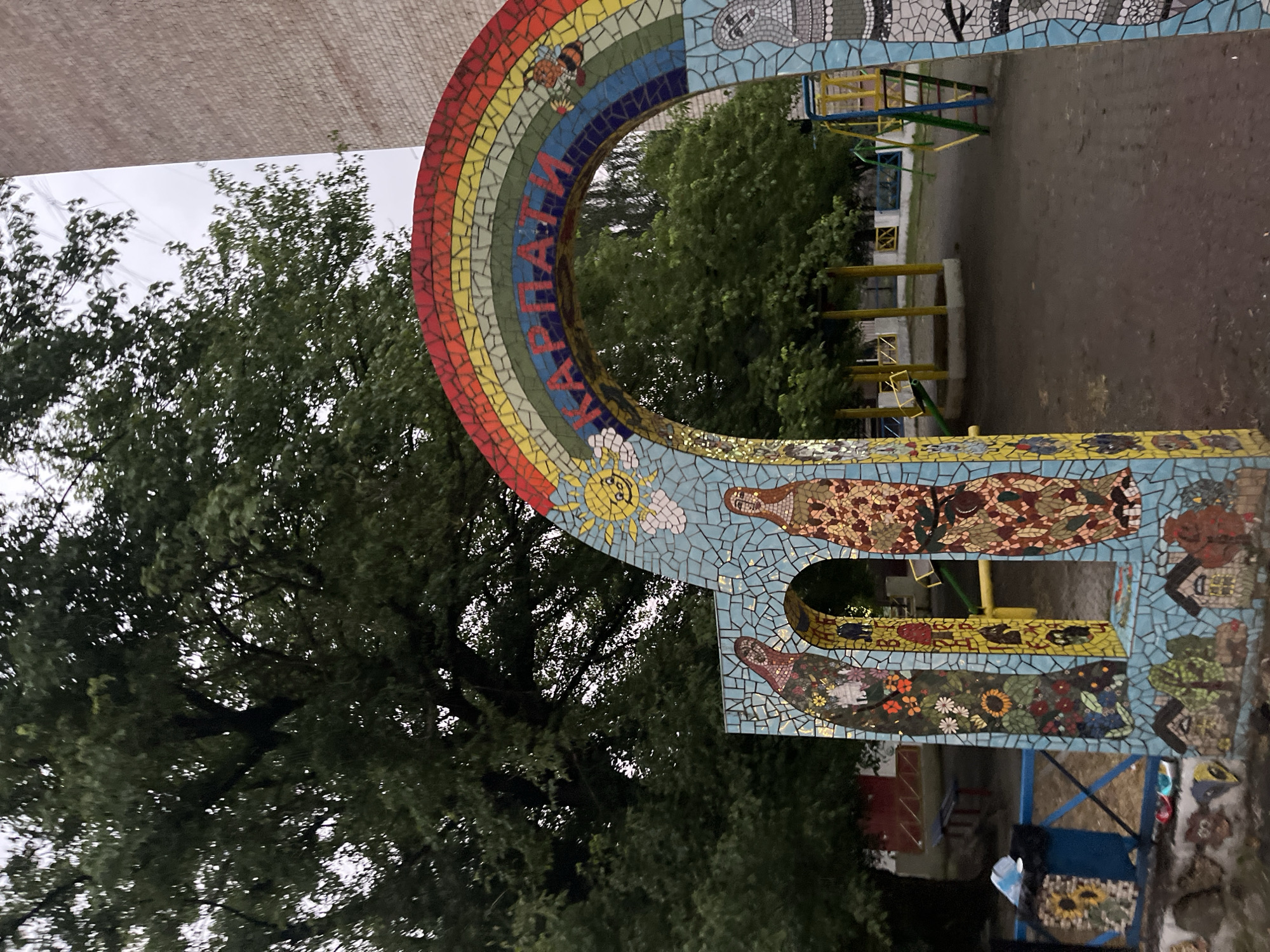 The image depicts an outdoor mosaic archway with the word "КАРПАТИ" (meaning "Carpathians") written across it in red lettering. The arch is adorned with a mosaic rainbow and various colorful mosaic designs, including a sun, a bee, and depictions of people in traditional clothing on the pillars. Below the arch, the mosaic continues with images of small houses and trees. The archway is situated in a playground area, evidenced by the presence of a slide and other play structures in the background. A large tree with lush green foliage is visible behind the archway, and a brick building stands to the right. A trash bin decorated with a sunflower mosaic is located to the left of the arch. The overall scene suggests a decorative entrance to a public space, possibly a kindergarten or recreational area, in Chernivtsi, Ukraine, given the Cyrillic inscription. The lighting suggests it is either overcast or late in the day. There are no people actively engaged in the scene.