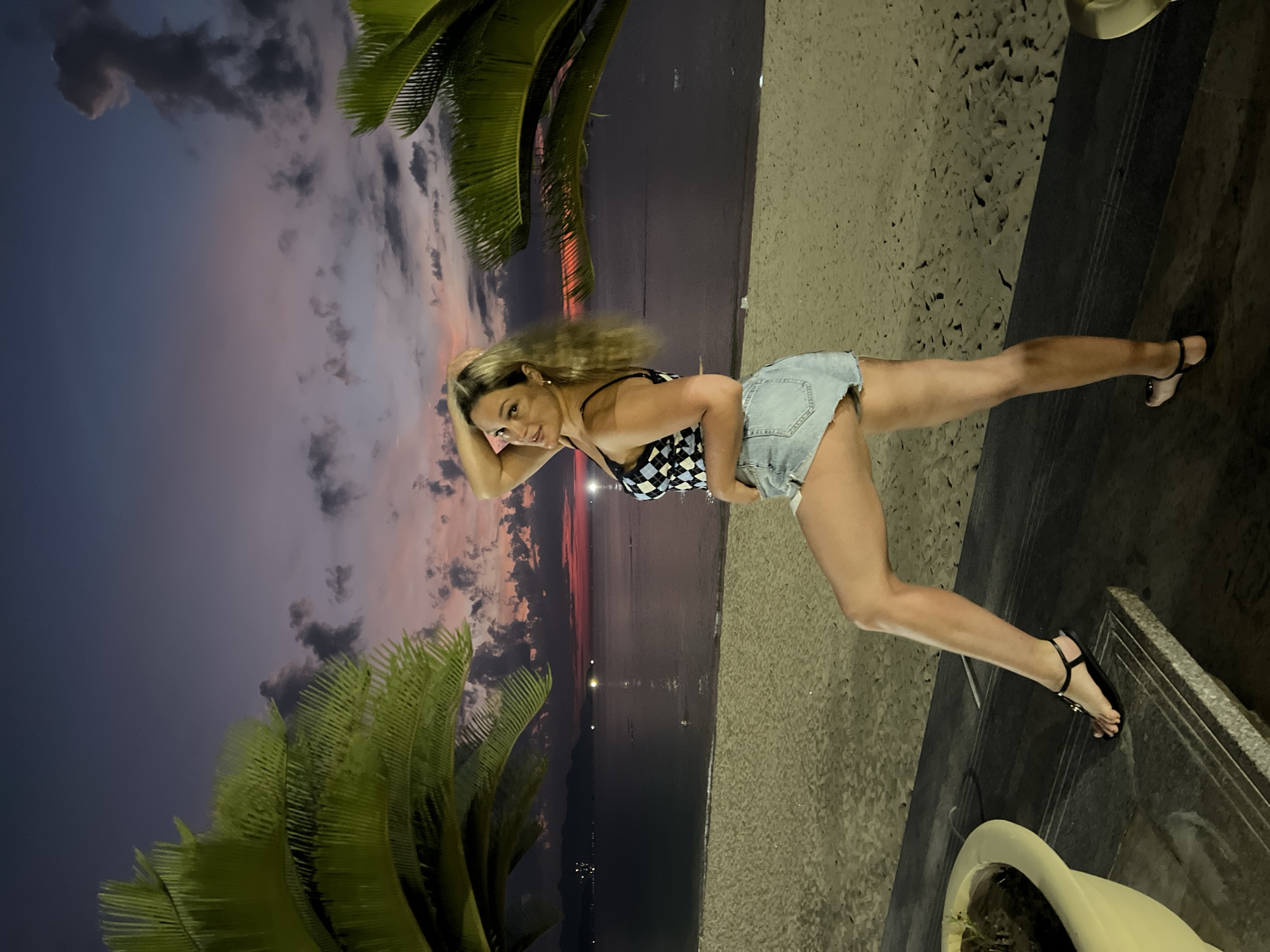 A woman is posing on a promenade adjacent to a beach in Nha Trang Ward, Vietnam at dusk. She is wearing a checkered top, denim shorts, and black sandals. She is positioned near a palm tree and a decorative planter. The background includes the beach, ocean, and a sky exhibiting colors of sunset. Artificial lighting is visible in the distance over the water.