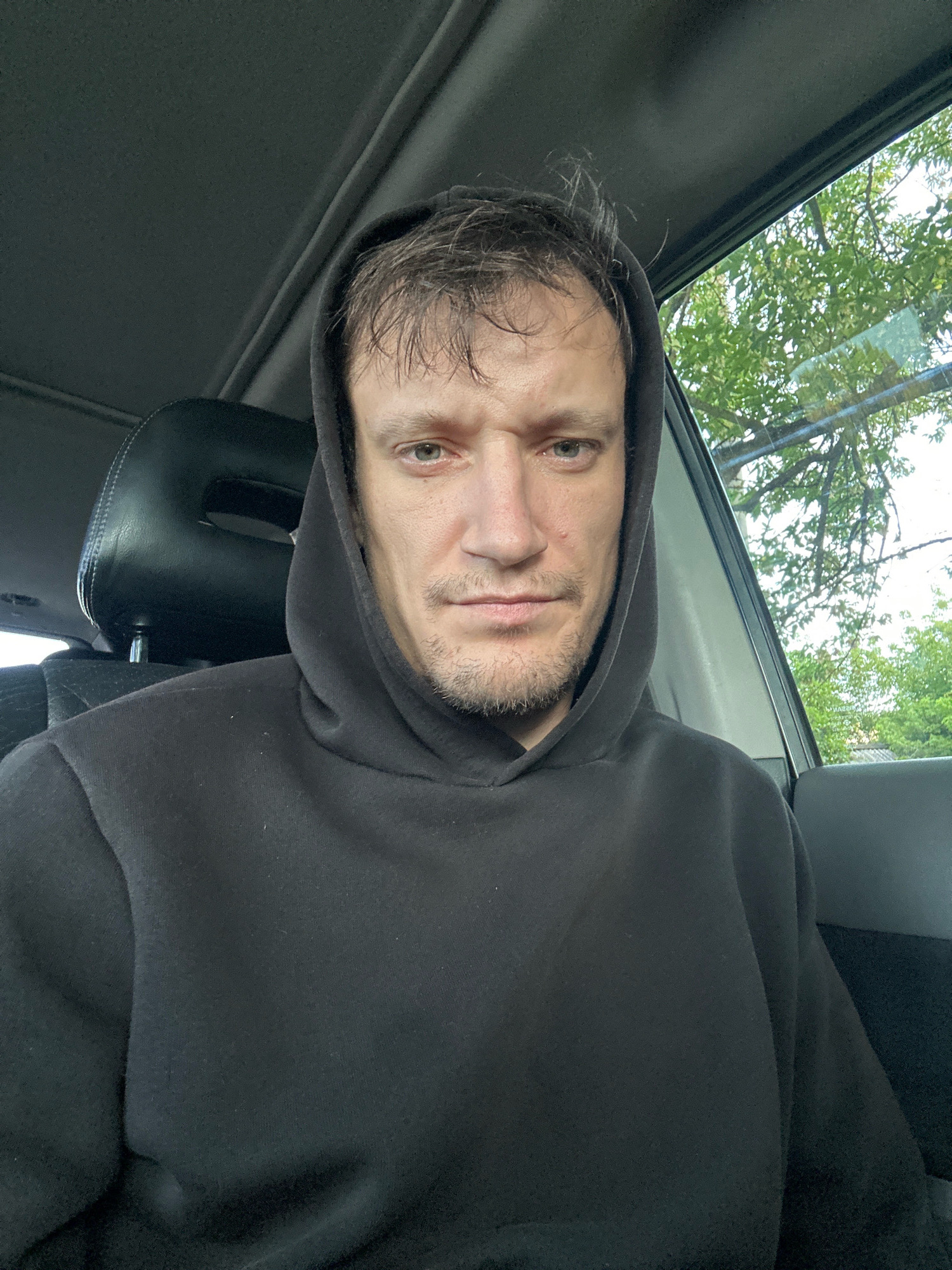 A selfie taken from inside a car shows a man wearing a black hoodie. He is looking directly at the camera, with his face slightly obscured by the hood. The man has light skin and short, dark hair that appears slightly disheveled. He has a faint stubble on his chin and a neutral expression. The interior of the car is visible behind him, including a headrest and part of the dashboard. The window to his right shows a blurry view of green trees and a cloudy sky, suggesting an outdoor setting in a city like Makiv, Ukraine.