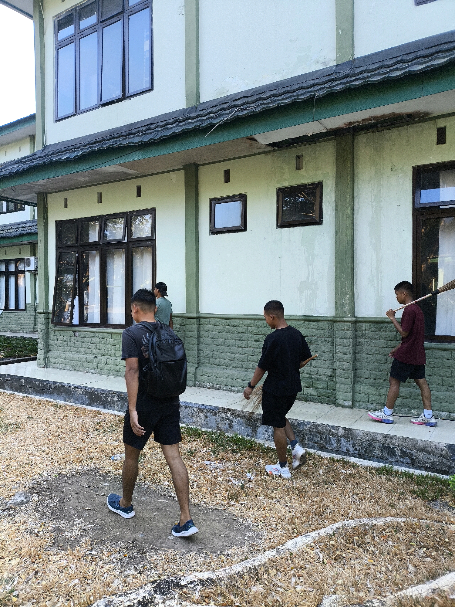 The image shows an outdoor scene in front of a two-story building with light green walls and dark green trim. The building has multiple windows with dark frames. In the foreground, three young men are walking along a paved path. The man on the left is walking away from the camera, wearing a black t-shirt, black shorts, and a black backpack. He is wearing blue slip-on shoes. The middle man is also walking away from the camera, wearing a black t-shirt and black shorts. He is wearing white athletic shoes with pink and blue accents. The man on the right is walking away from the camera, carrying a broom over his right shoulder. He is wearing a maroon t-shirt and black shorts. He is wearing white athletic shoes with pink and blue accents. The ground is covered with dry leaves and some patches of grass. Another man is visible in the background, standing near a window. The scene appears to be in a public space, possibly a school or administrative complex, given the building's architecture. The overall impression is one of routine activity, with the men seemingly engaged in some task or movement along the path.