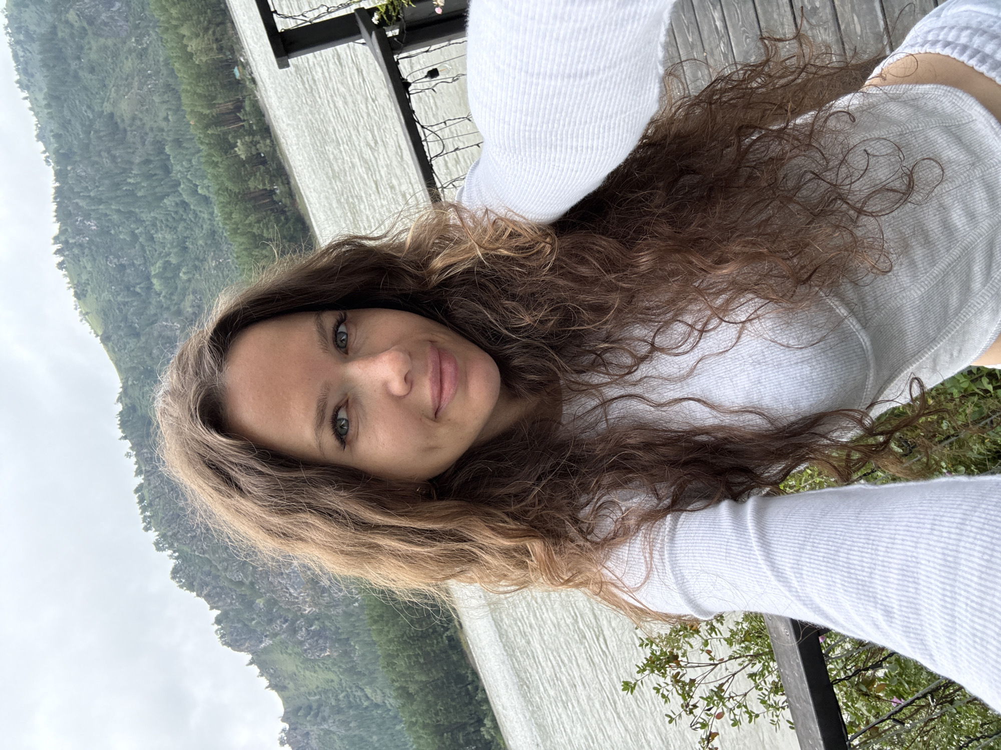 A young woman with long, curly brown hair is taking a selfie outdoors. She is wearing a white long-sleeved shirt. The background shows a body of water, a forested mountain range, and a wooden deck. The lighting is overcast. The location is stated to be Nizhny Kayancha, Russia.