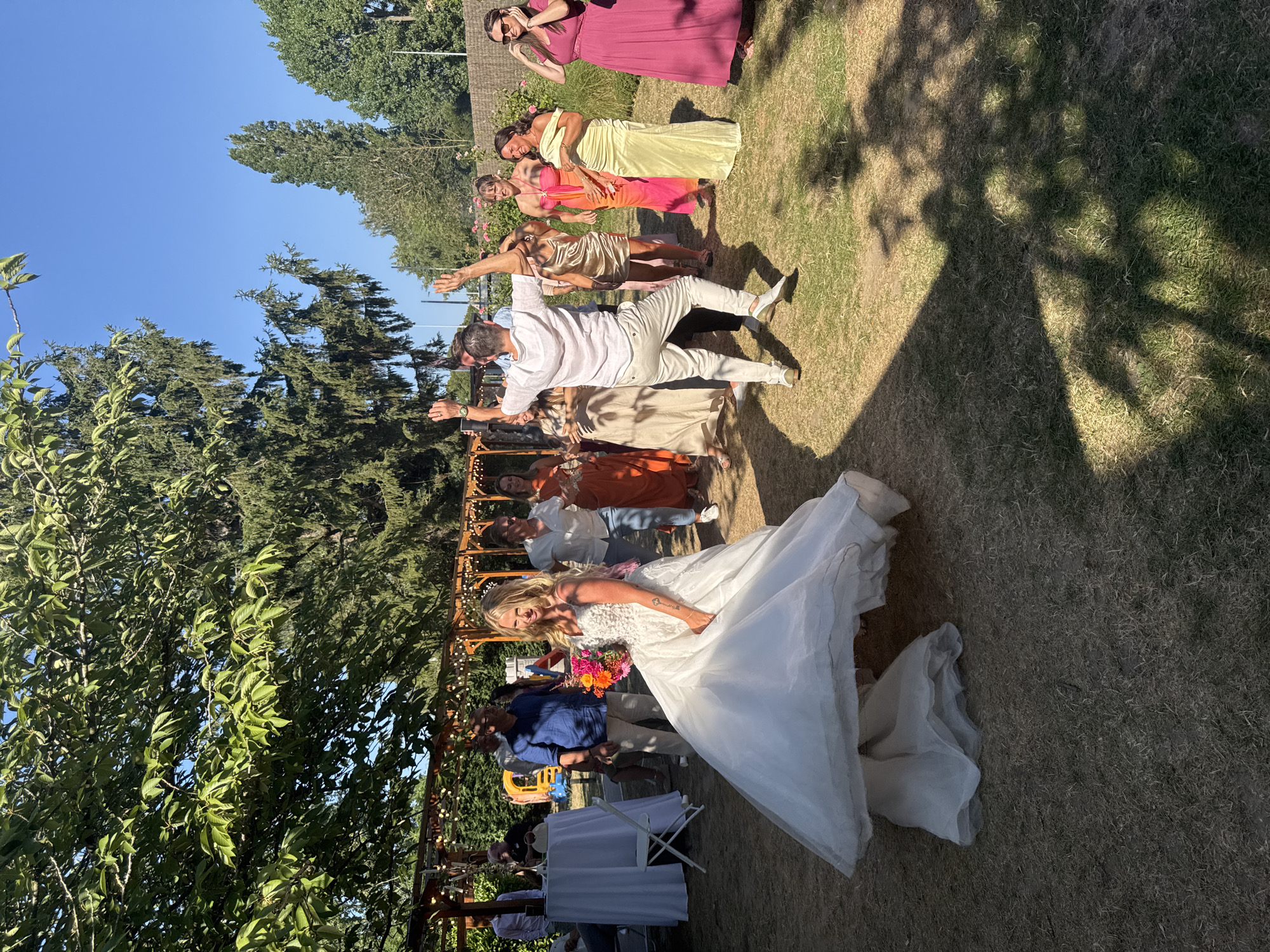 In Hem, France, a wedding celebration unfolds outdoors on a sunny day. A bride in a white wedding gown, her dress billowing around her as she moves, is captured in motion. She holds a colorful bouquet of flowers. Beside her, a man in a white shirt and beige pants dances with his arms raised. A group of guests in various colorful dresses and outfits watch and engage with the celebration. In the background, trees and a clear blue sky form the backdrop, with string lights and a wooden pergola suggesting a decorated outdoor venue. The scene is set on a lawn with scattered dry patches of grass.
