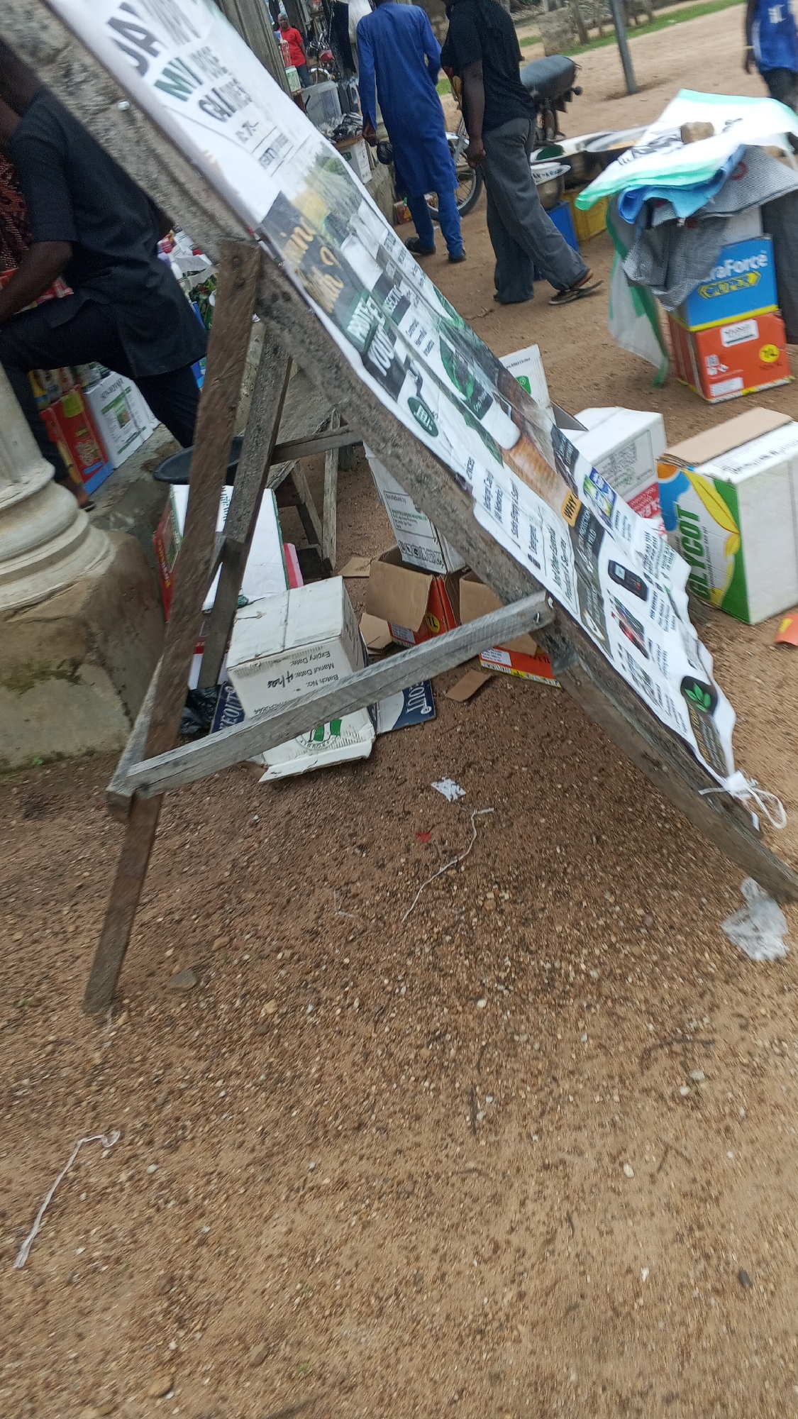 The image depicts an outdoor scene in Nyita, Nigeria, characterized by a dirt and gravel ground. A wooden A-frame stand displays a large poster or banner covered in text and images, possibly advertising or informational. The poster appears to be wrapped around a cylindrical object, suggesting it might be a roll-up banner or a large cylindrical advertisement. Several cardboard boxes are scattered around the base of the stand, some open and others stacked. One prominent box in the foreground is white with black text, and another box to the right is blue and orange with "AuraForce" branding. In the background, several individuals are present. One person is wearing a blue robe, another a black shirt and grey pants, and another a black shirt. A motorcycle is also visible in the background. The overall impression is of a market or roadside stall environment.