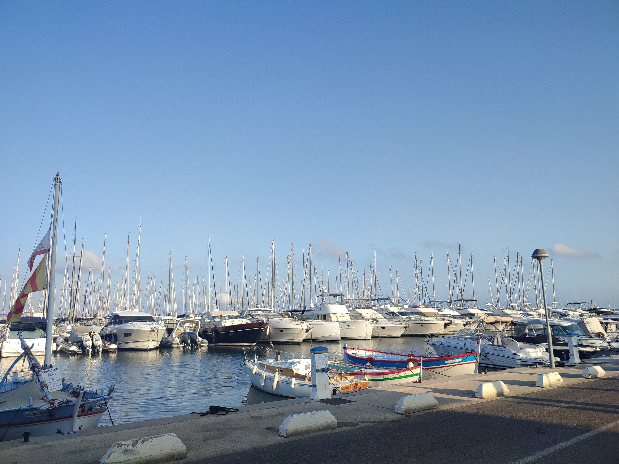 The image depicts a marina in Hyères, France, likely on a clear, sunny day. Numerous sailboats and motorboats are moored in the water, filling the frame from left to right. The boats range in size, with a prominent white sailboat in the foreground on the left and several larger yachts and smaller vessels behind it. Some boats have sails furled, while others appear ready for departure.

Along the waterfront, a concrete promenade is visible with a few white, rectangular bollards. A lamppost stands near the right side of the image. The water of the marina is calm and reflects the blue sky and the surrounding boats. In the background, the sky is a clear, vibrant blue with a few wispy clouds near the horizon. The overall impression is one of a peaceful and well-maintained harbor. There are no people visible in the image.