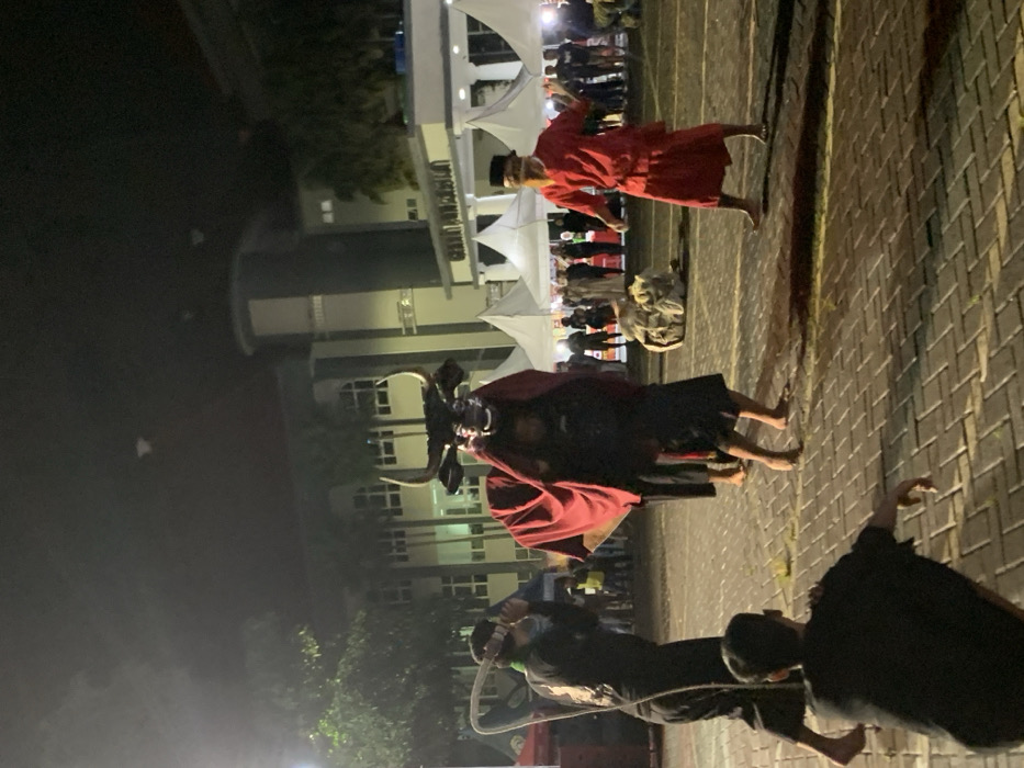 The image captures a performance in an outdoor area in Kecamatan Batu, Indonesia, located at latitude -7.867 and longitude 112.514. The performance appears to be a traditional Indonesian dance, possibly a re-enactment of a mythical event. A person dressed in black and red clothing is carrying a large black bull's head mask on their shoulders, with red fabric draped over the mask and down to the floor. The person is walking towards the right of the image. To the left of the dancer, a man dressed in black is holding a rope and watching the dance. Another man, wearing a black shirt and pants, is sitting on the ground watching the performance. In the background, a large building with a white awning is visible, with several white tents set up in front of it. Several people are standing around the tents, watching the performance. The stage is illuminated by artificial lighting, casting a warm glow on the performers and audience.