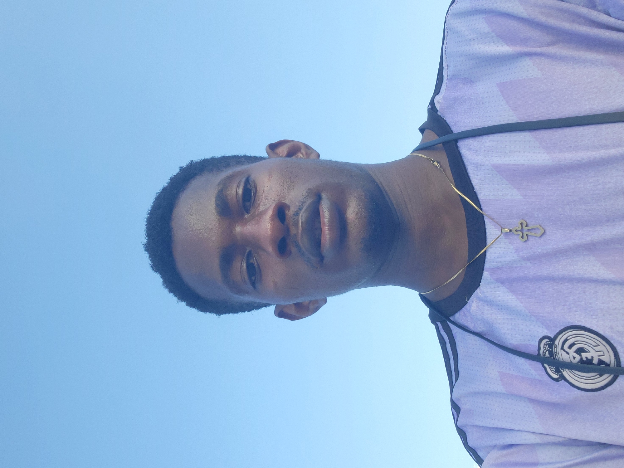 This is a low-angle, portrait-style photograph taken outdoors on a clear, sunny day. The subject is a young Black man, seen from the chest up, facing the camera. He has short, dark, curly hair and is wearing a light purple and white jersey with dark stripes on the shoulders. A thin gold chain with a cross pendant is visible around his neck. The background is a solid, clear blue sky, suggesting it is daytime. The image has a somewhat casual, selfie-like feel, with the subject's face close to the camera. The man appears to be looking directly at the viewer, with a neutral expression. The location is identified as Egbagu, Nigeria.